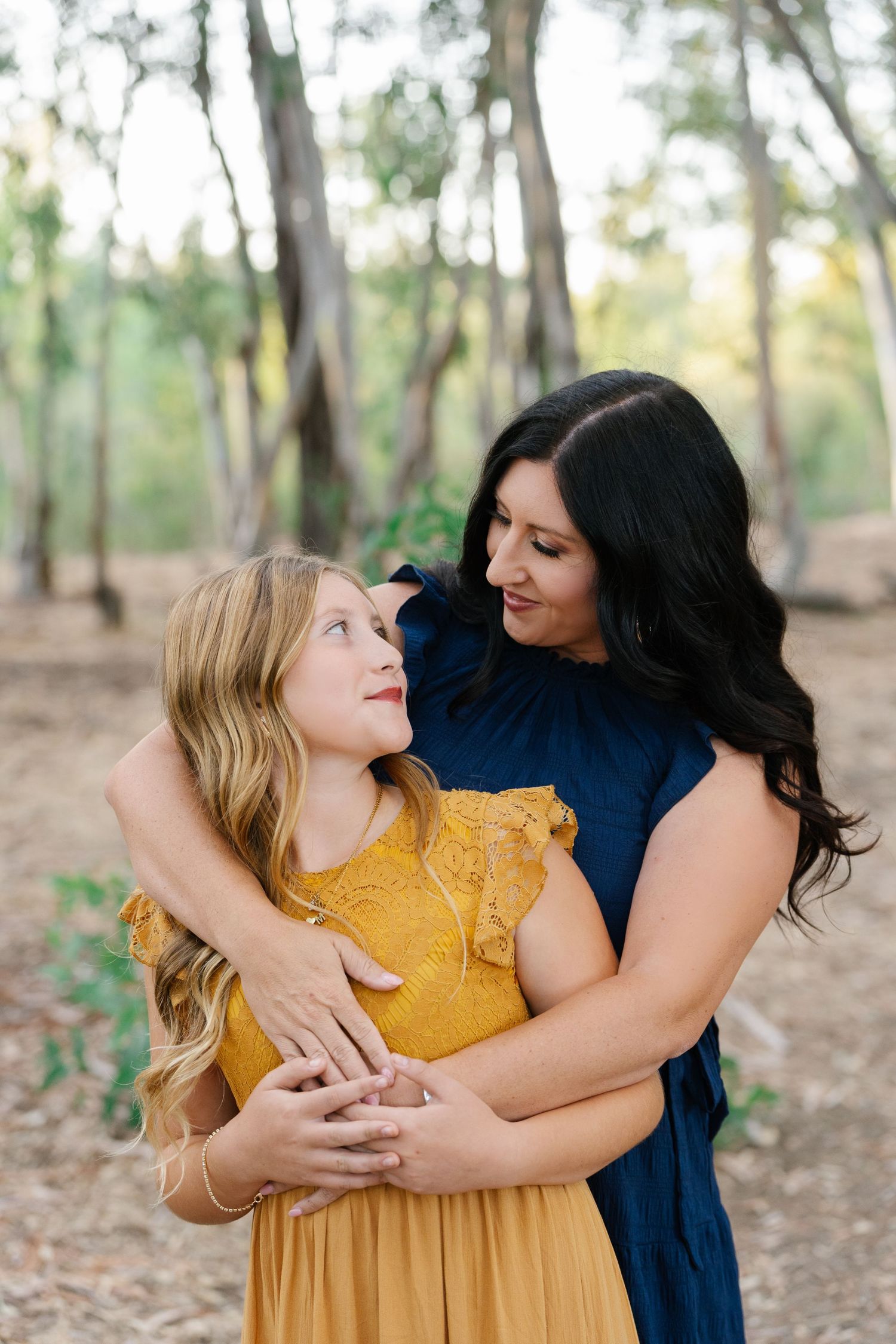 A mother and daughter share a warm embrace in a wooded setting, wearing navy and mustard dresses.
