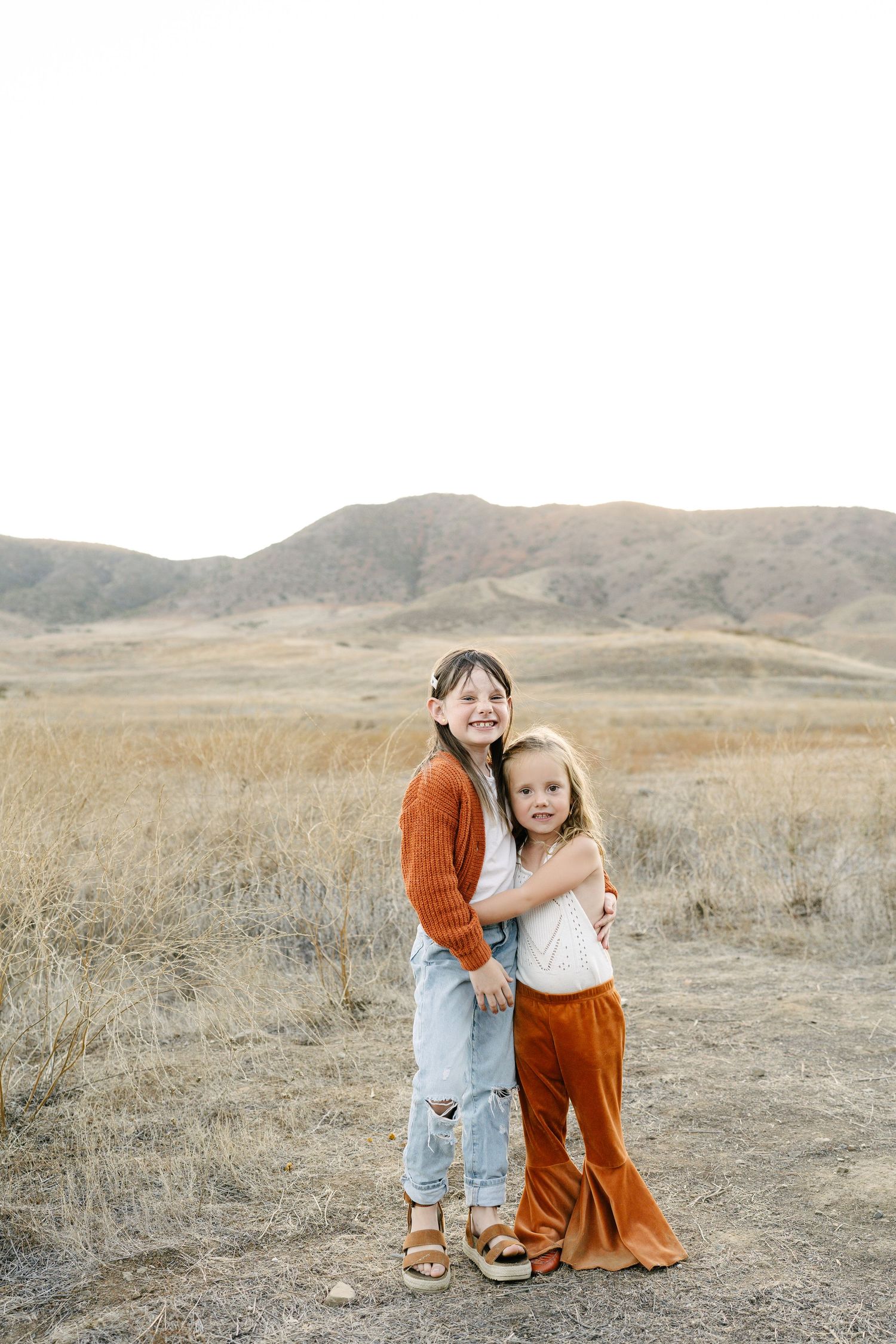 Two young sisters hug each other in a dry golden field with rolling hills behind.