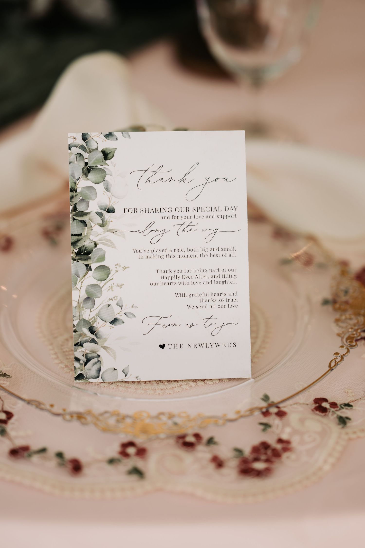 Elegant wedding thank you card with eucalyptus greenery border displayed on a decorative plate at a reception table.