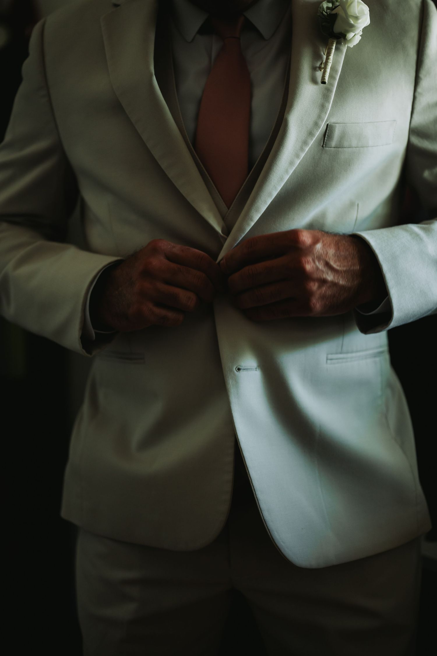 Groom in white suit with red tie and boutonniere buttoning jacket before wedding ceremony.