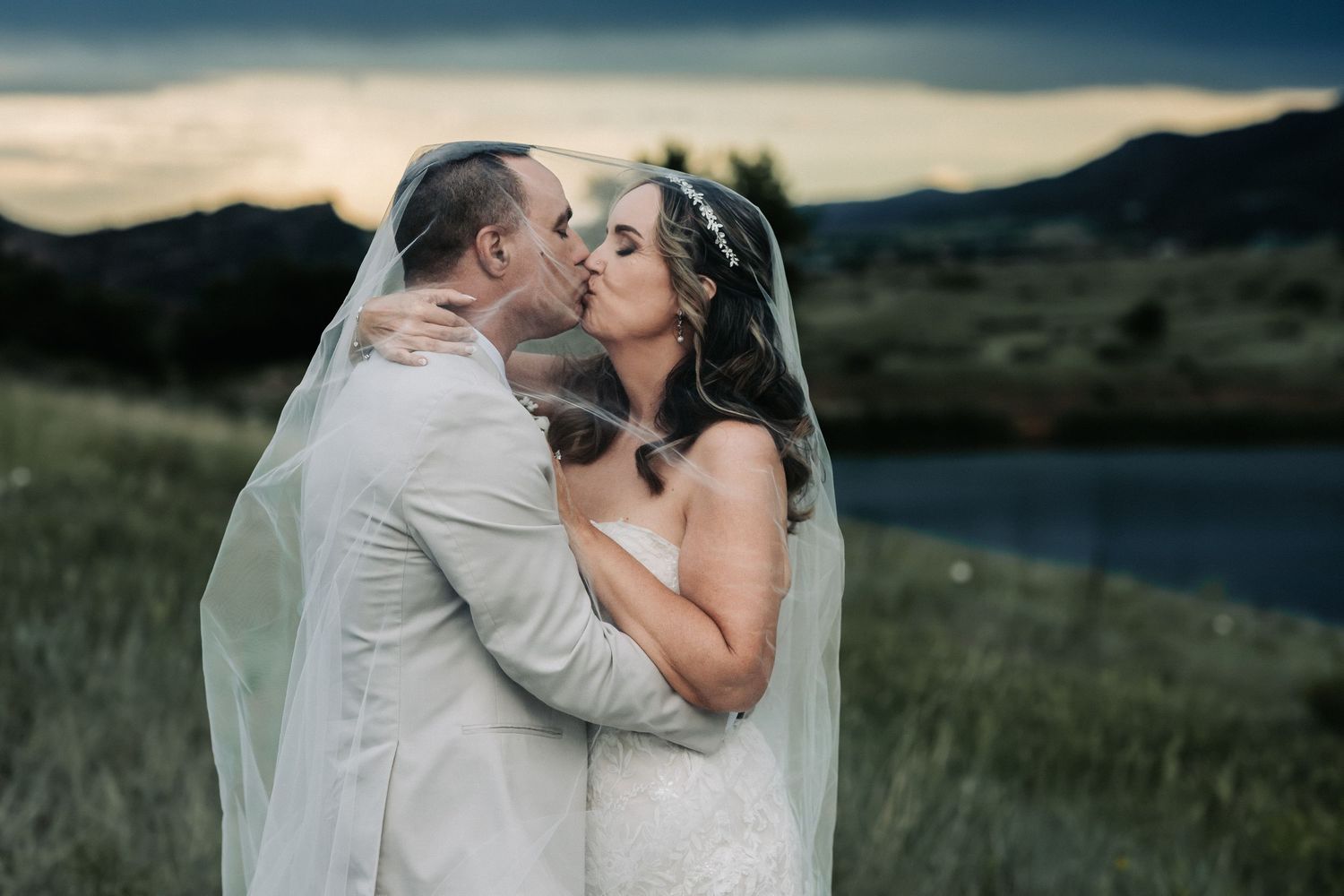 Bride and groom share a romantic kiss at sunset, with mountains and a lake in the background.
