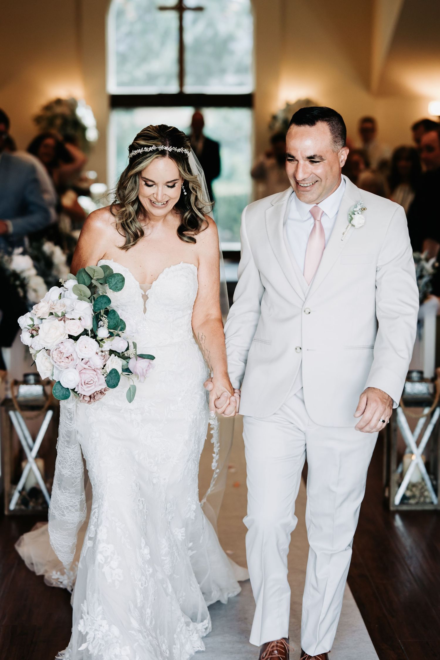 Bride in lace gown and groom in ivory suit walk down the aisle holding hands after wedding ceremony.
