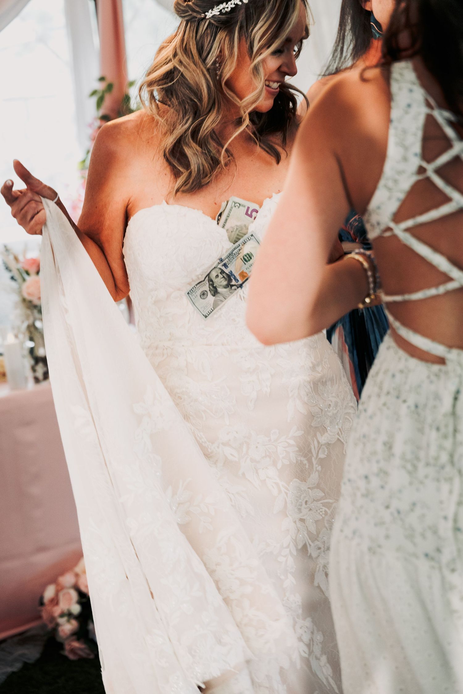 Bride in lace wedding dress smiling as bridesmaid helps tuck money into her strapless gown.