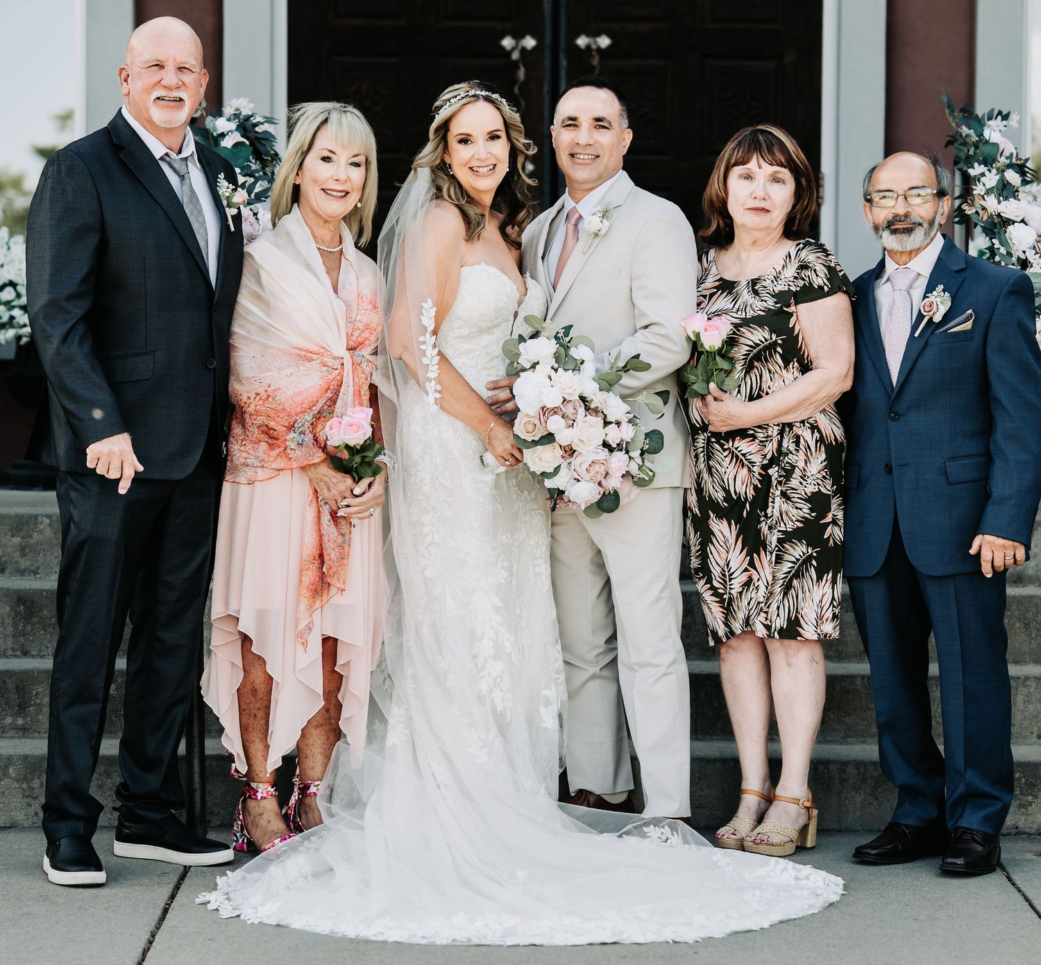 Wedding couple poses with family members outside a church, bride in white gown holding pink bouquet.