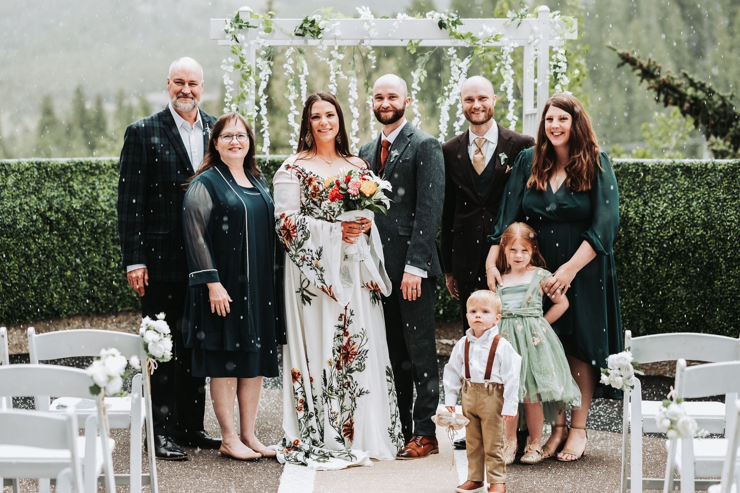 Wedding party poses outdoors at elegant venue with floral arch, bride in floral gown with bouquet, surrounded by family.