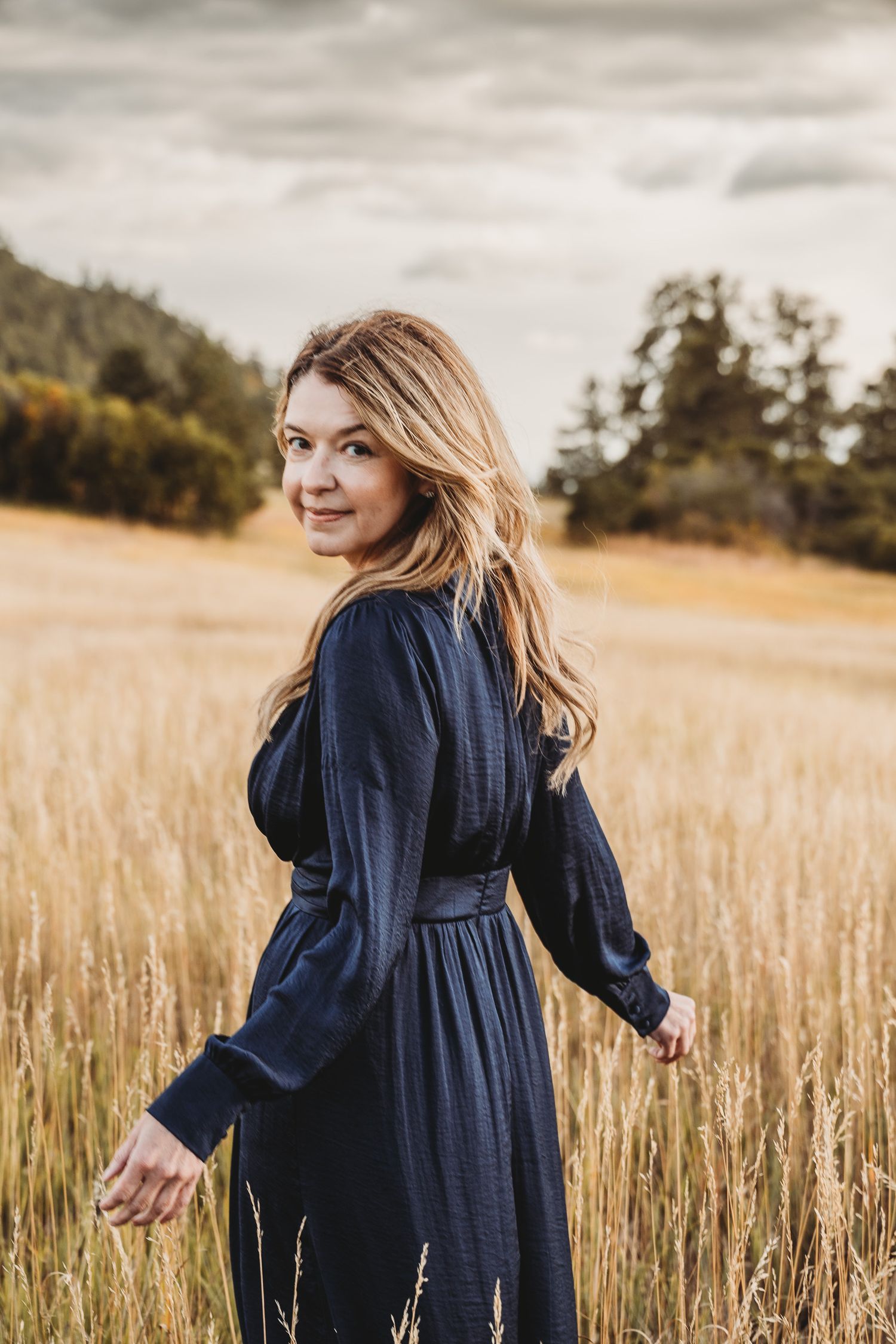 Person in navy blue dress walks through golden wheat field at sunset in artistic lifestyle photoshoot.