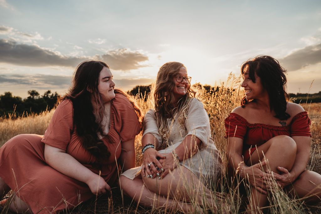 A group of friends in rust-colored dresses lay together in a golden wheat field at sunset.