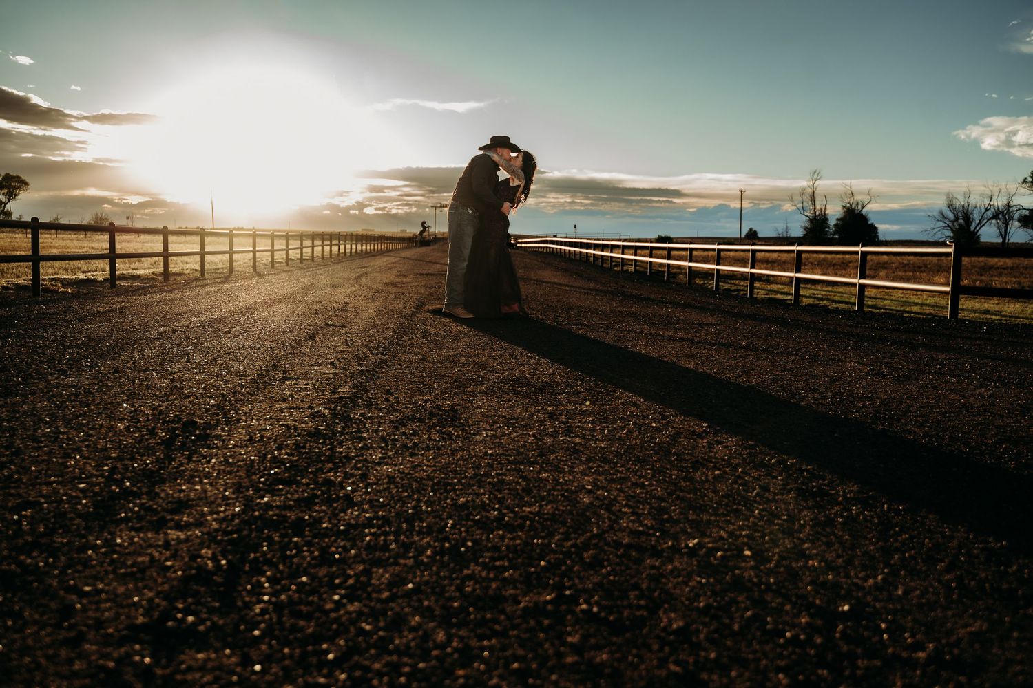 Couple embracing in a silhouette kiss on a rustic dirt road lined with wooden fences as the sun sets over open farmland.