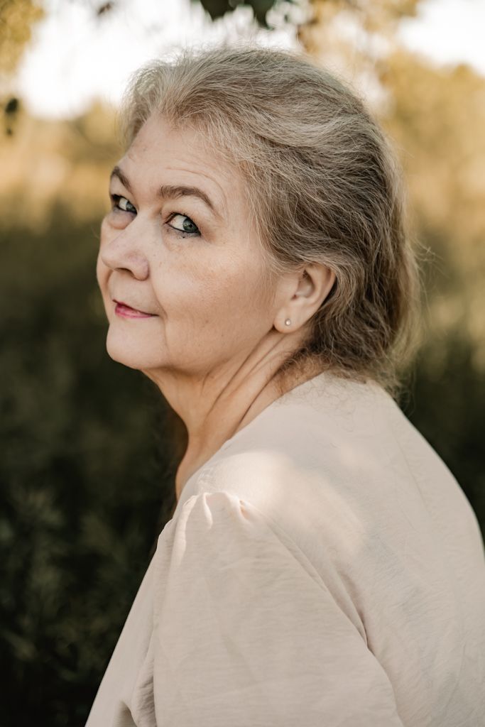 Portrait in soft lighting showing a gentle smile and cream-colored top against a blurred natural background.