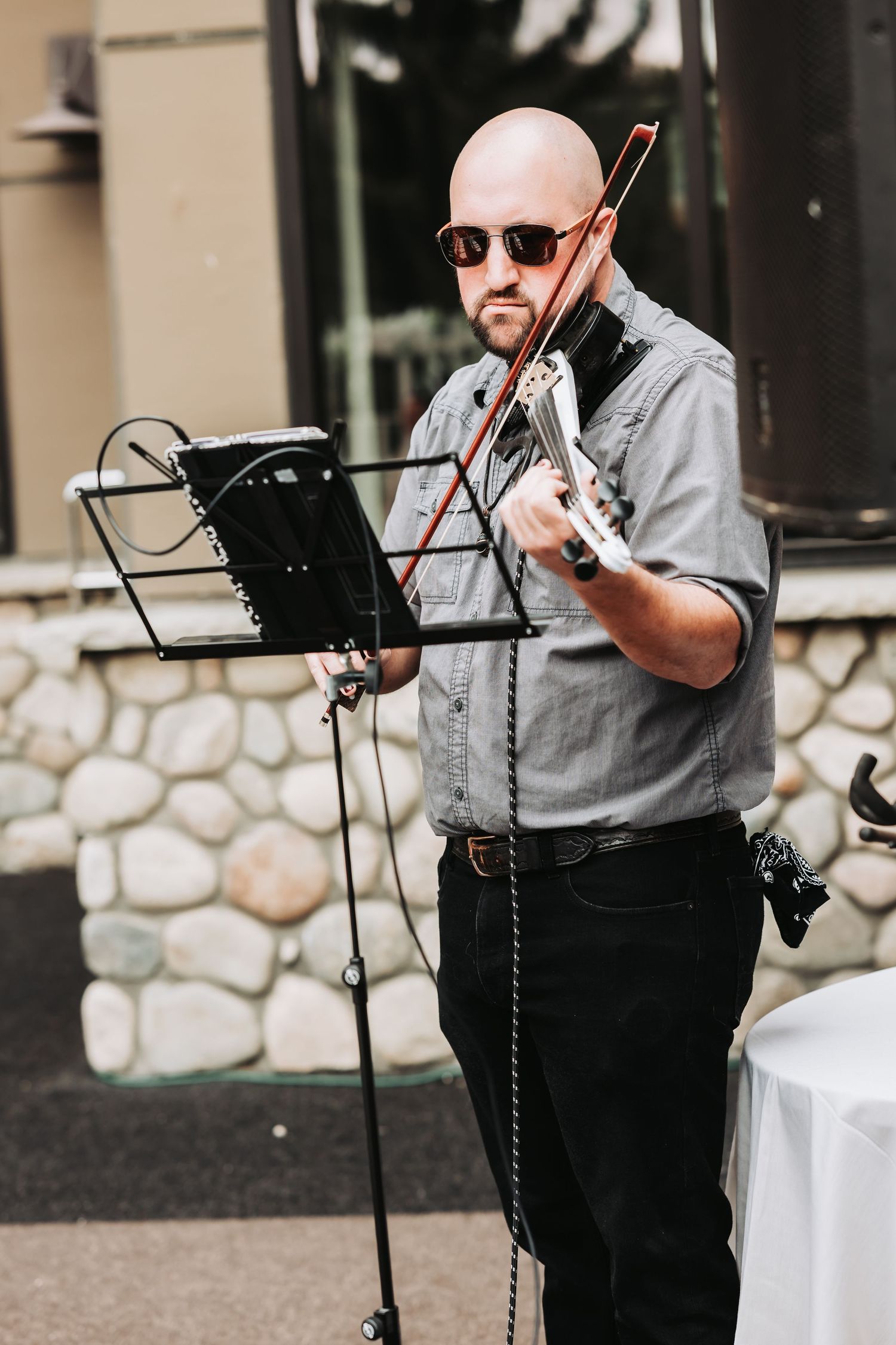 Bald musician in sunglasses plays electric violin outdoors at a music stand near a stone wall.
