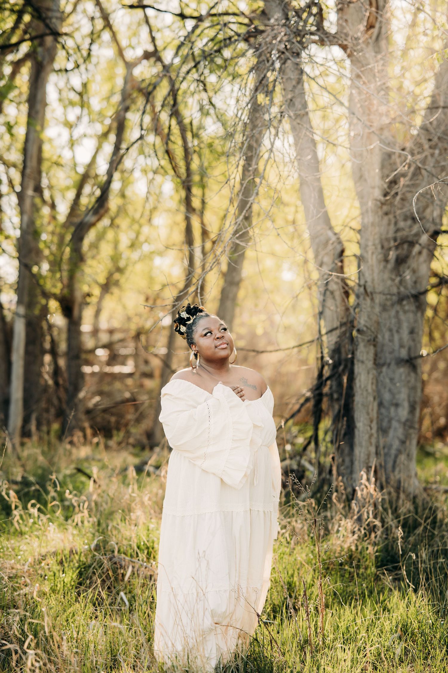 A smiling woman in a flowing white off-shoulder dress stands among sunlit trees in a natural woodland setting.
