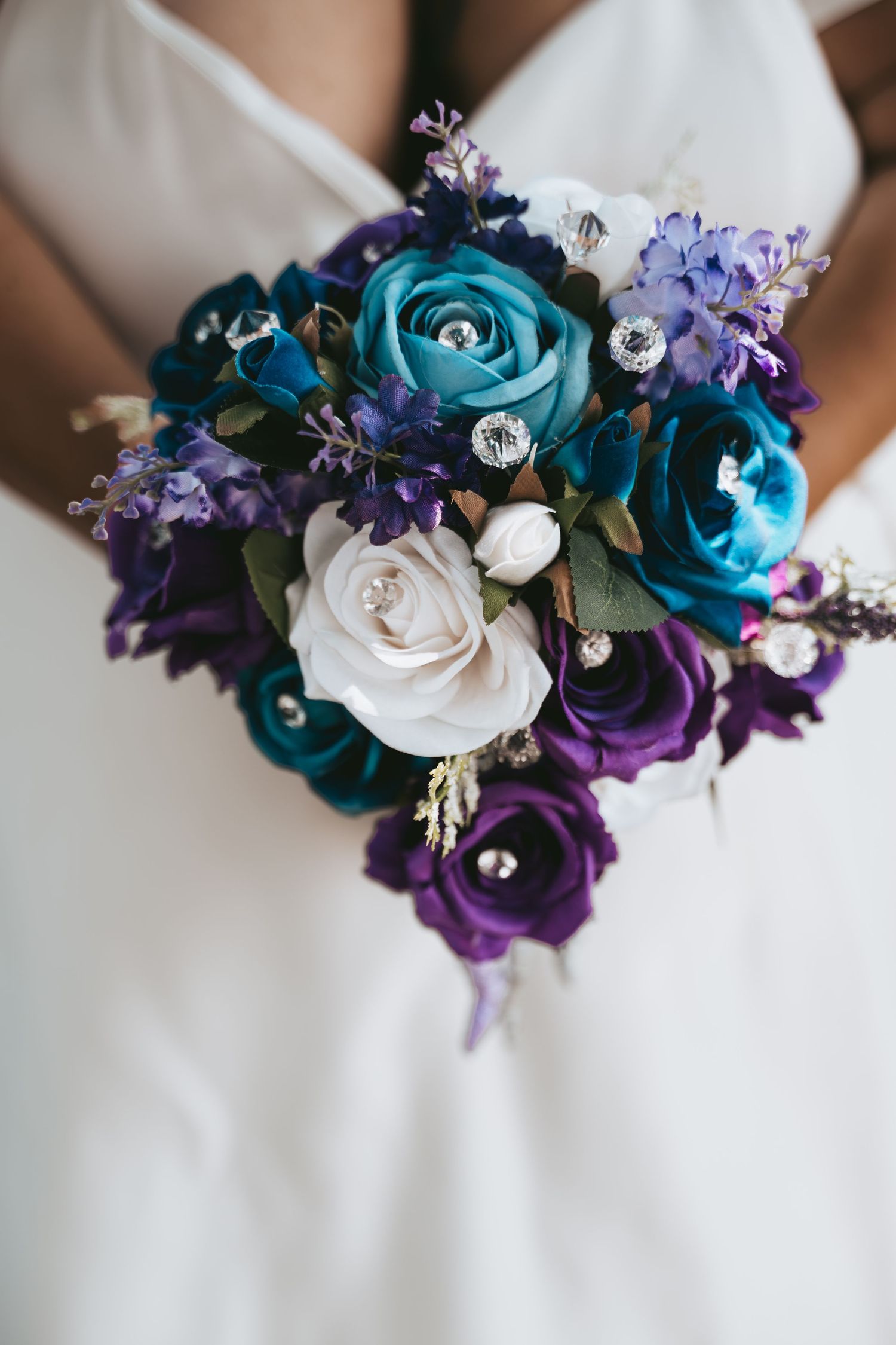 Bride holding a vibrant wedding bouquet with teal, purple, and white roses accented with crystal brooches.