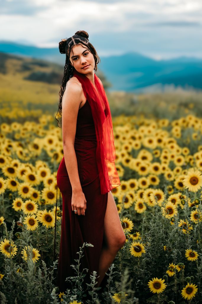 A person in a burgundy dress poses gracefully among vibrant yellow sunflowers in a scenic mountain field.