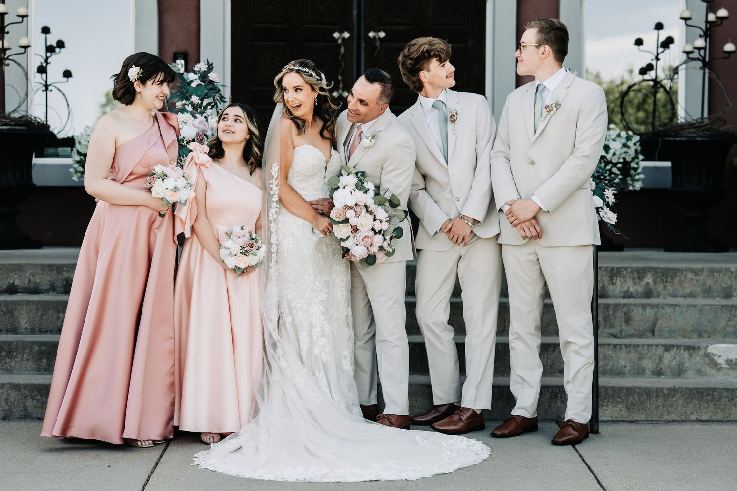 Bride and groom pose with wedding party in blush pink bridesmaid dresses and tan suits outside a venue.