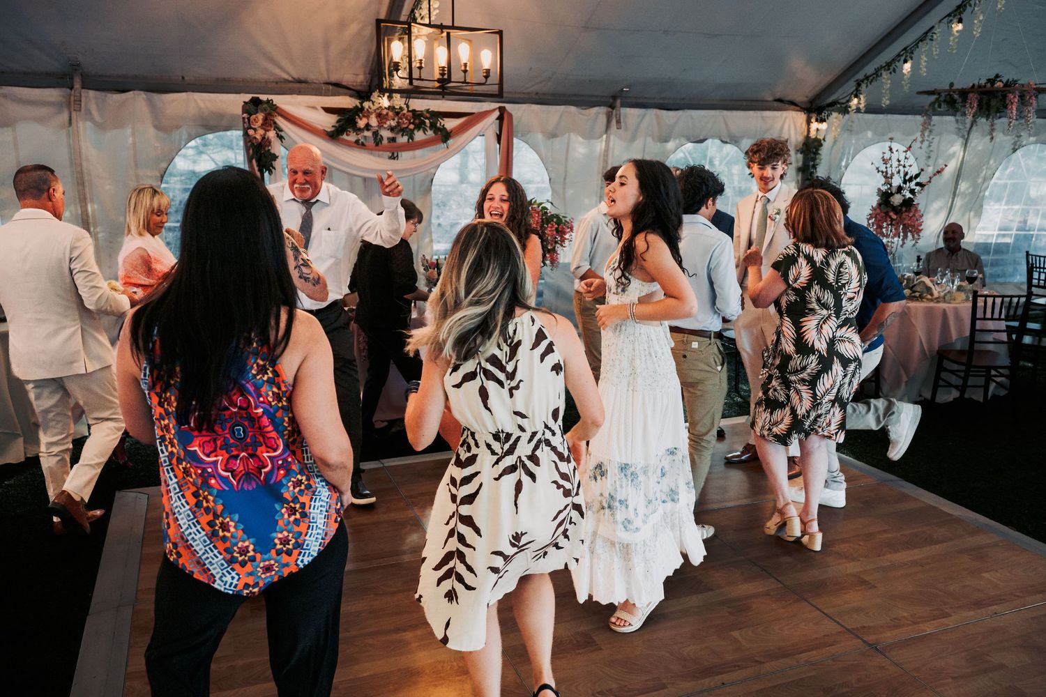 Guests dancing and celebrating at an elegant outdoor wedding reception under a white tent with chandelier lighting.