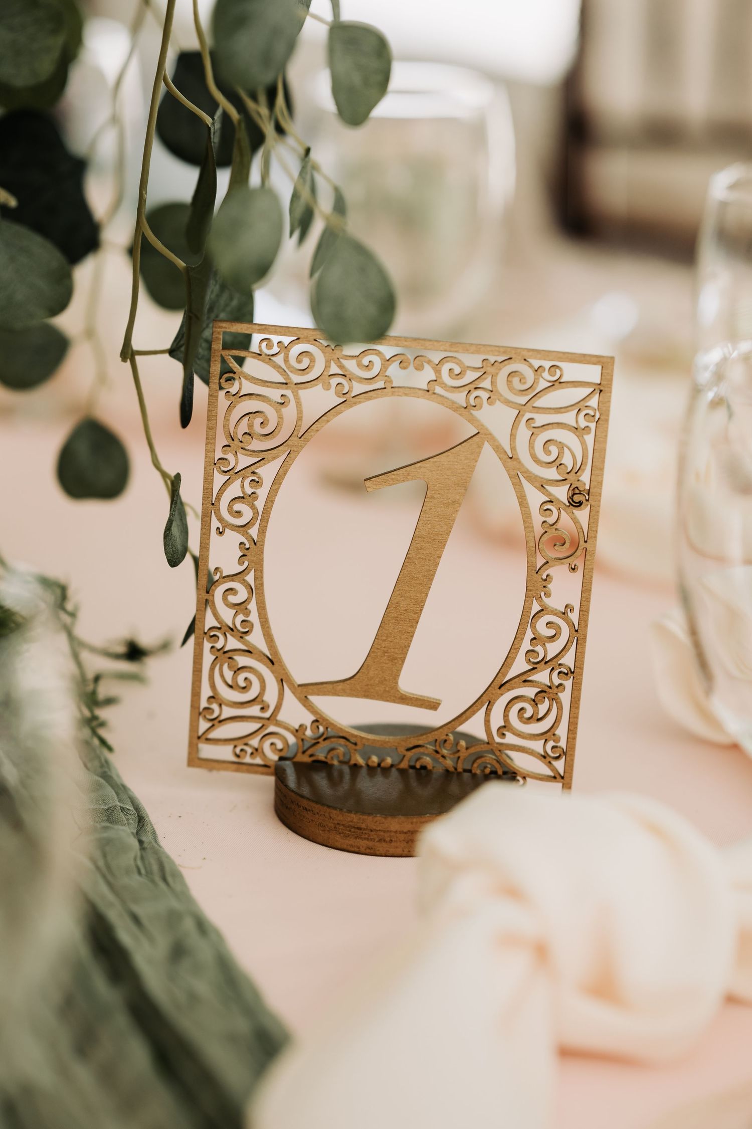 Gold laser-cut filigree table number one on wooden stand with eucalyptus greenery at elegant wedding reception.