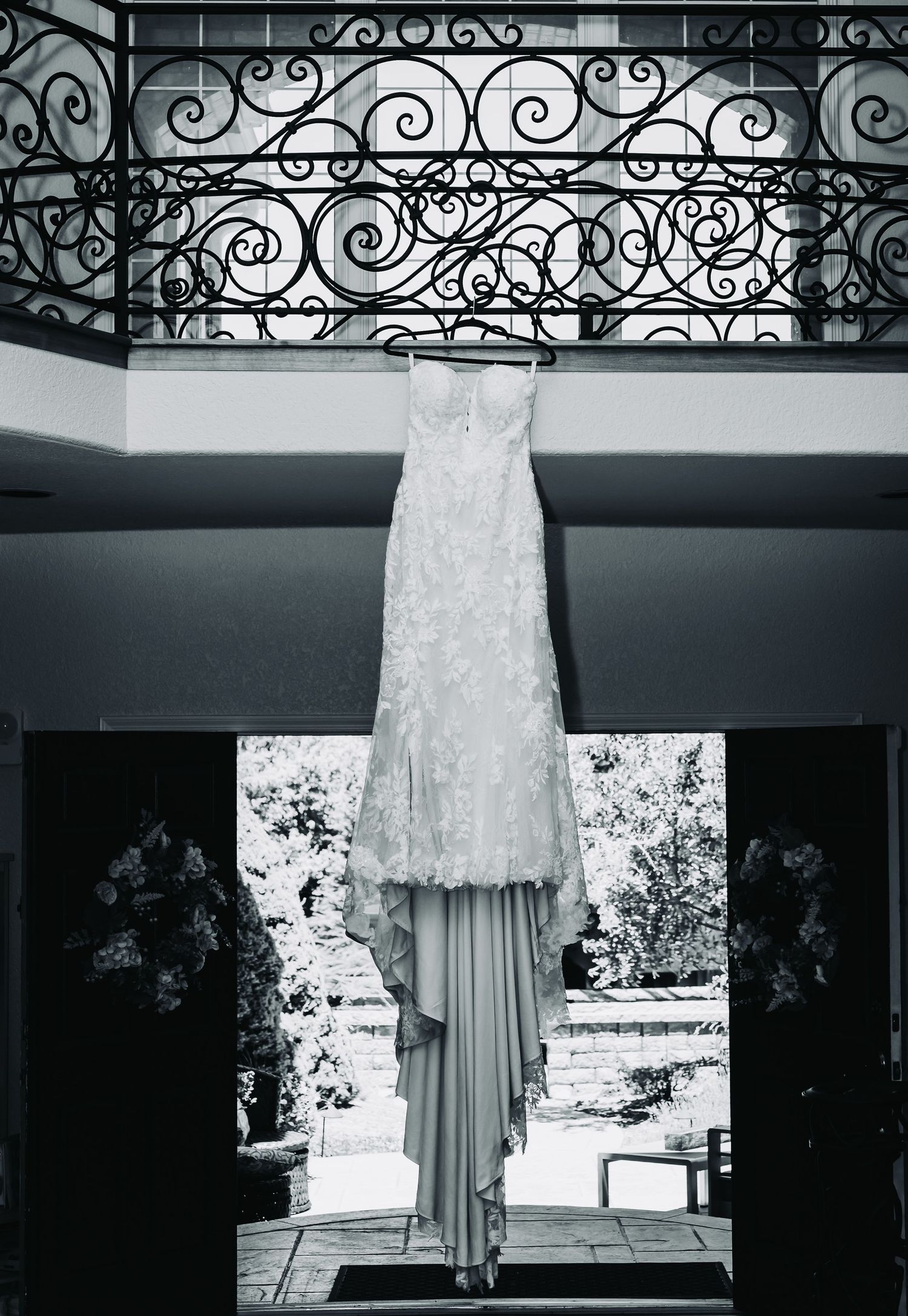 Black and white photo of an elegant lace wedding dress hanging on a hanger in front of ornate iron balcony railing.