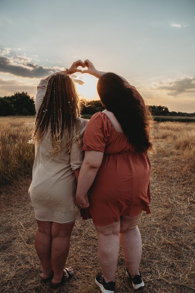 Two friends in casual attire stand together in a golden field at sunset making heart shapes with their hands.