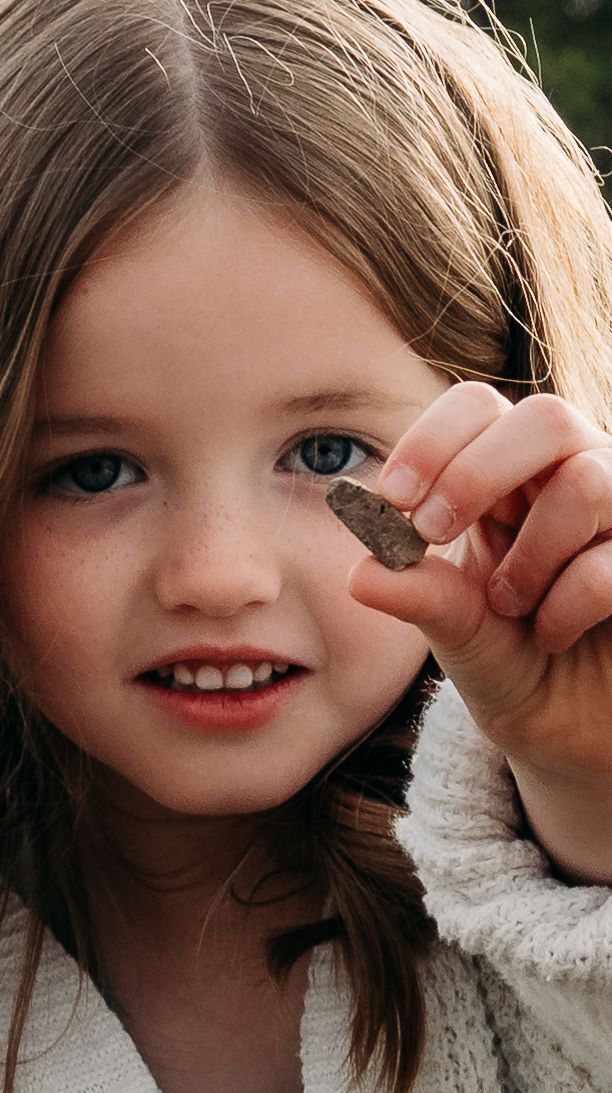A young girl with blue eyes smiles while holding a small rock up to the camera outdoors.