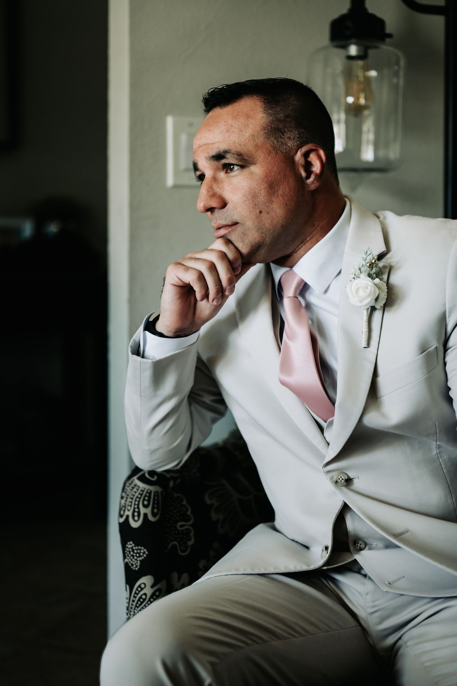 A contemplative groom in a white suit with pink tie and boutonniere sits pensively, resting his chin on his hand.