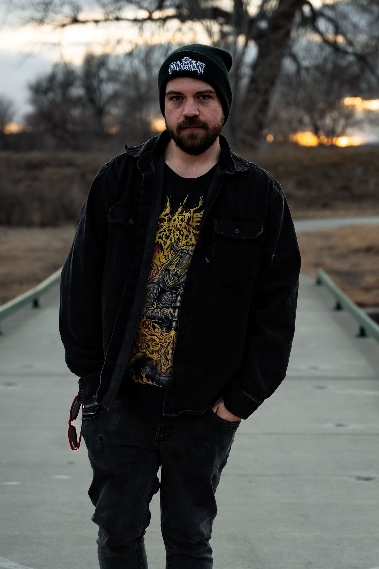 Person in black beanie and jacket stands on concrete path at dusk with dramatic sky behind.