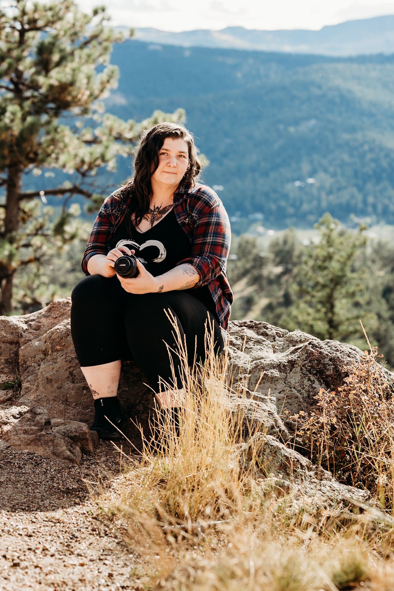 Woman sitting on rocky outcrop holding camera with mountain forest backdrop on sunny day.