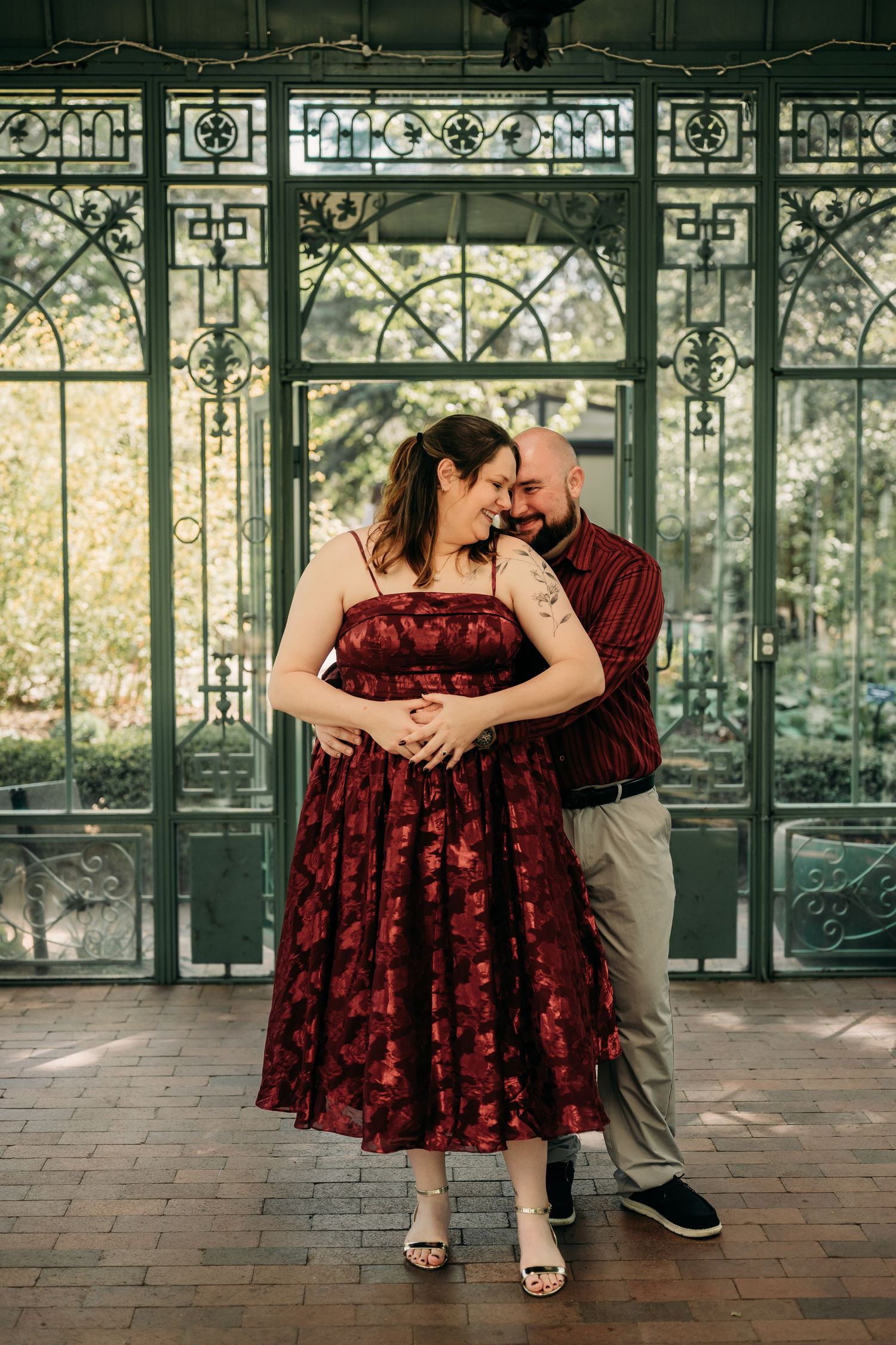 Couple in matching burgundy outfits embracing and laughing in front of ornate green iron greenhouse doors.