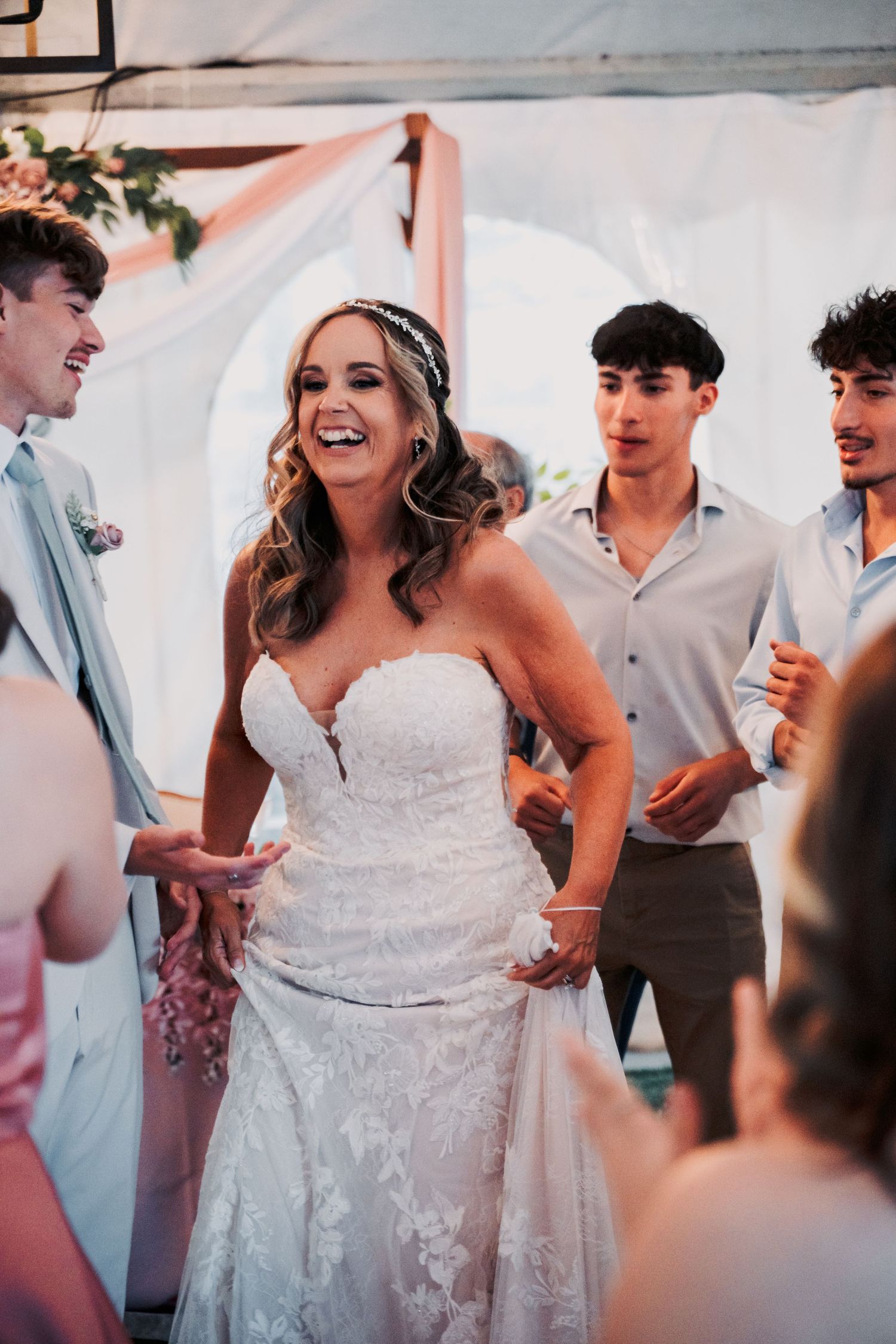 Joyful bride in lace strapless wedding gown laughing with guests during wedding reception dance.