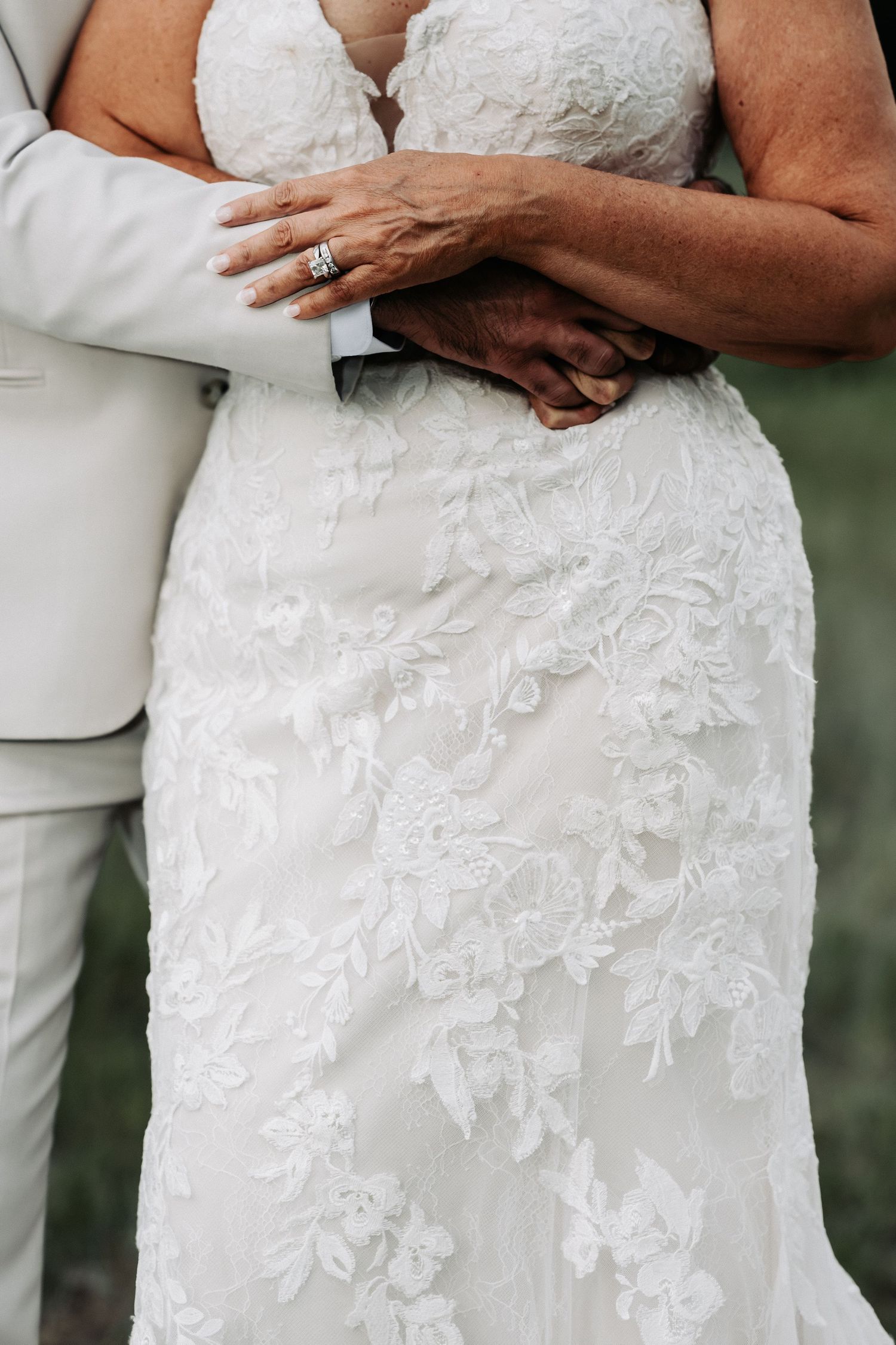 Close-up of bride in lace wedding gown and groom in gray suit holding hands, showcasing diamond engagement ring.