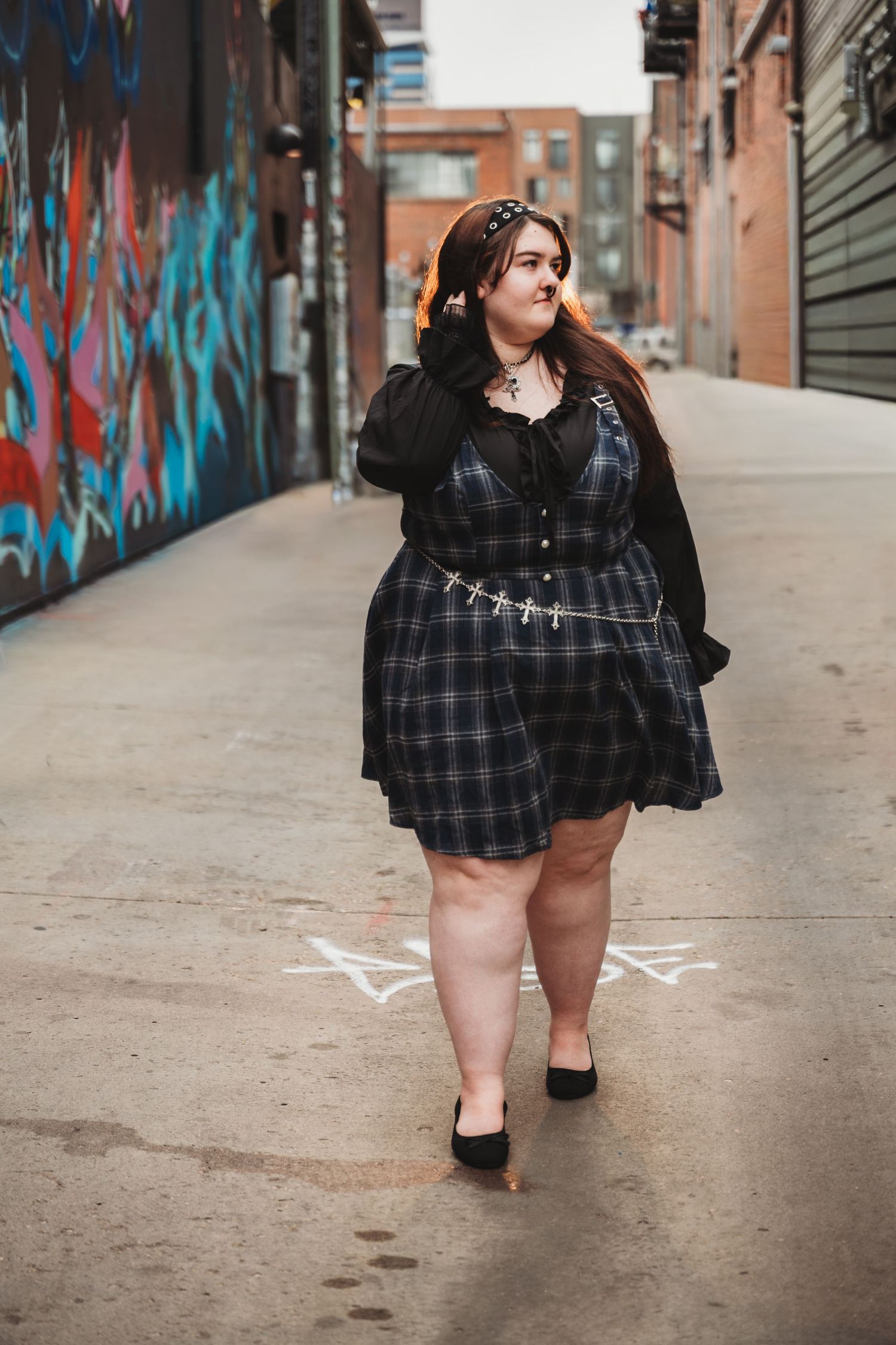 Plus size fashion model in plaid dress poses confidently against urban graffiti wall backdrop.