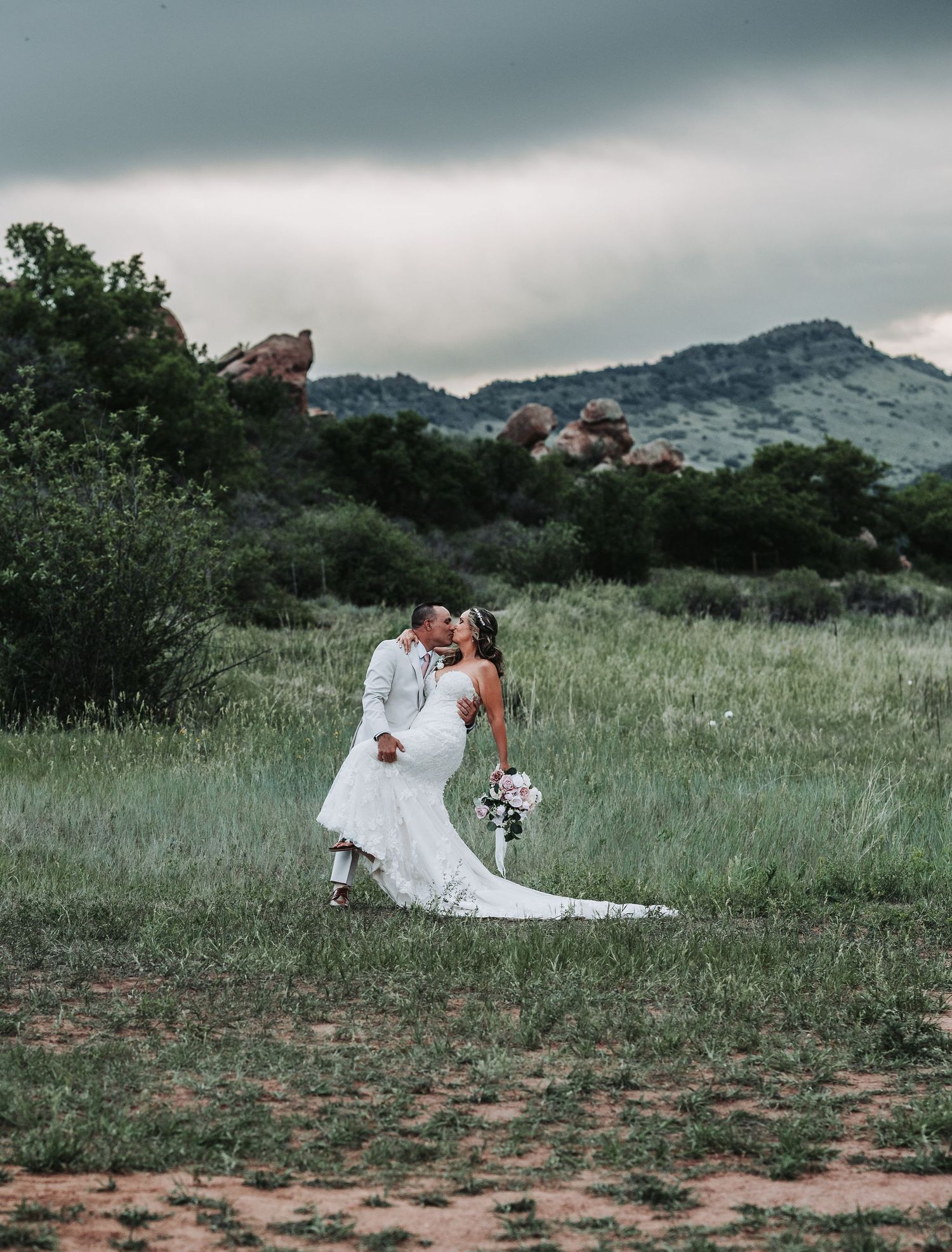 A bride and groom share a romantic dip kiss in a lush green meadow with rocky mountains under a dramatic cloudy sky.