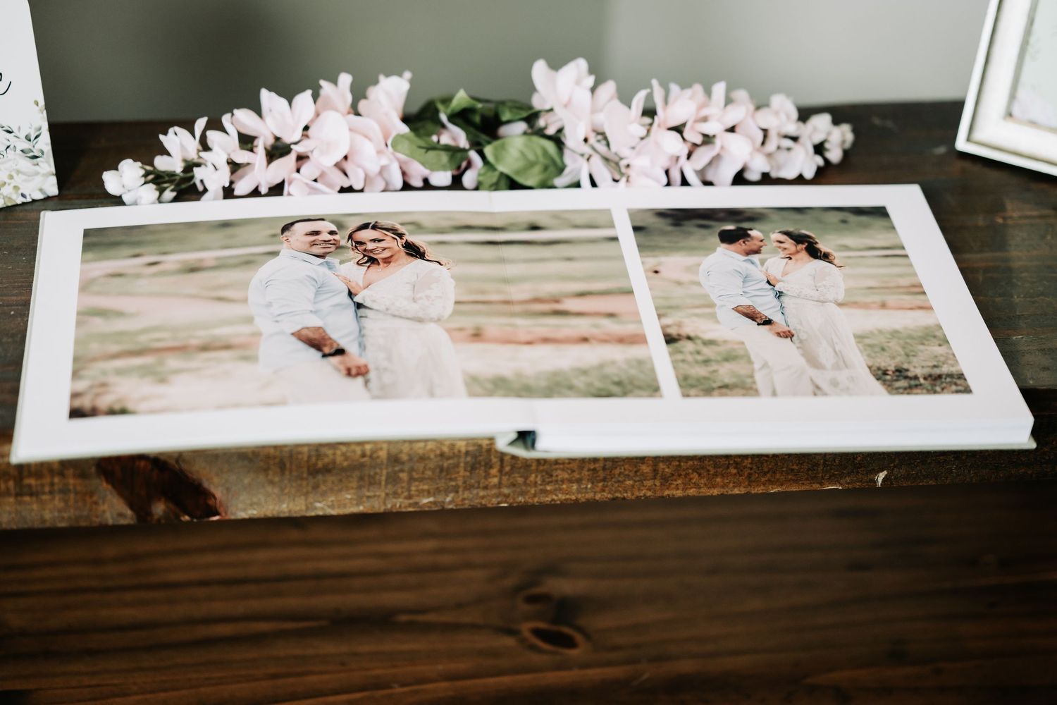 A wedding photo album open on a wooden table displaying romantic couple portraits with floral decorations in the background.