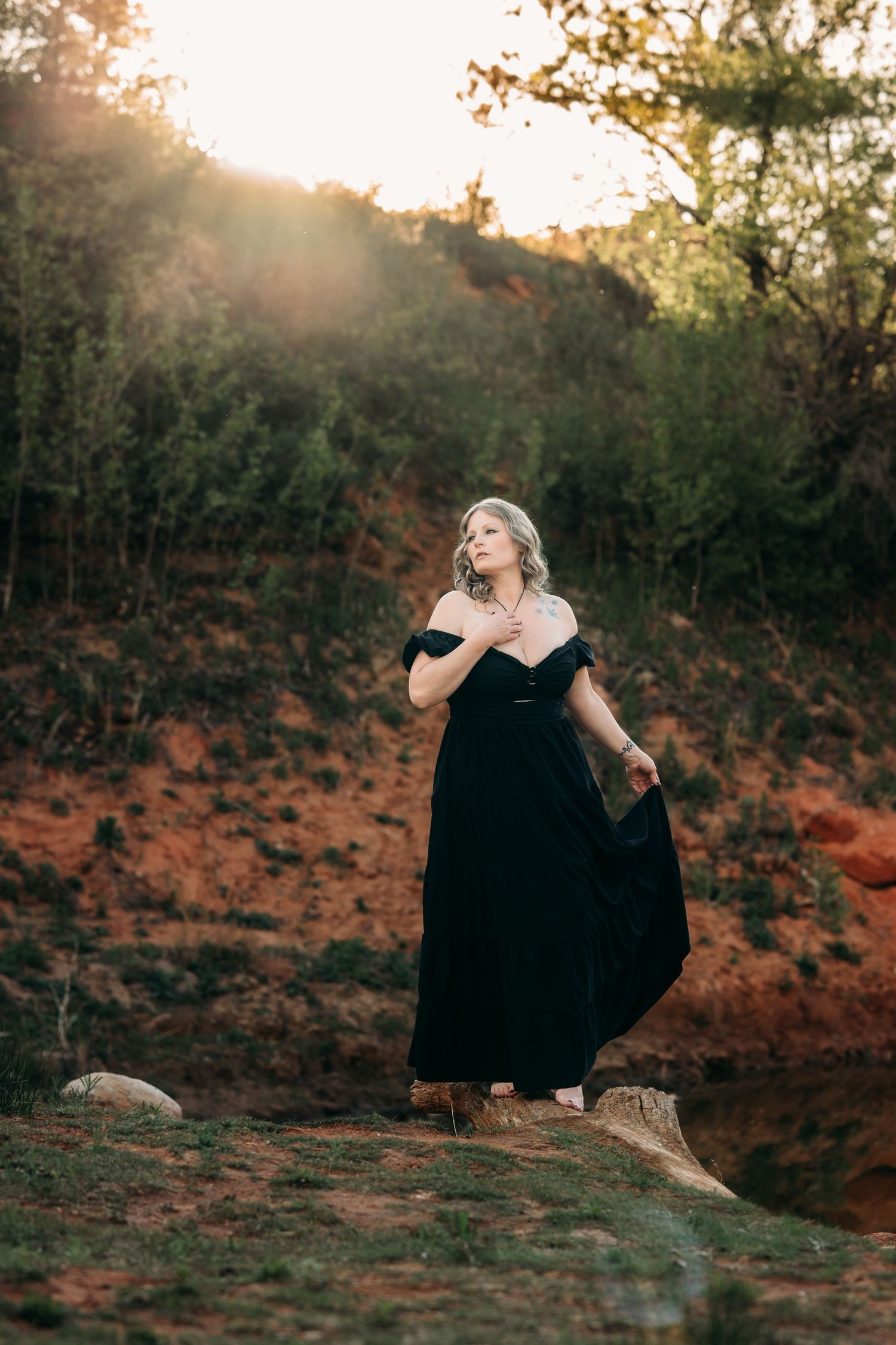 Woman in black off-shoulder gown poses outdoors at golden hour with red earth and green trees behind her.