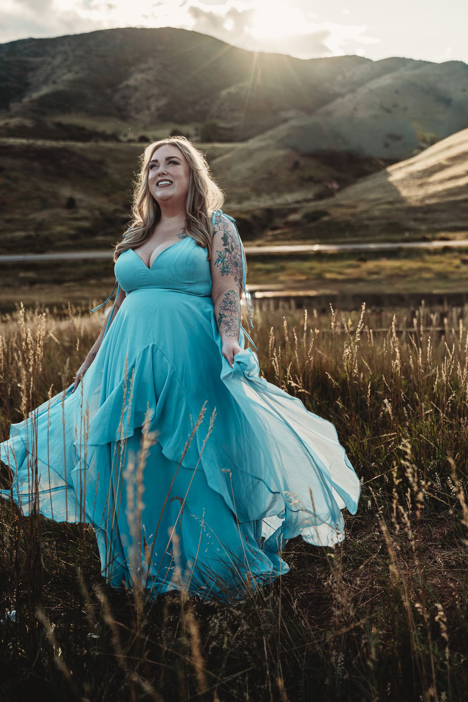 Woman in flowing teal gown poses in golden field with sunlit hills backdrop.