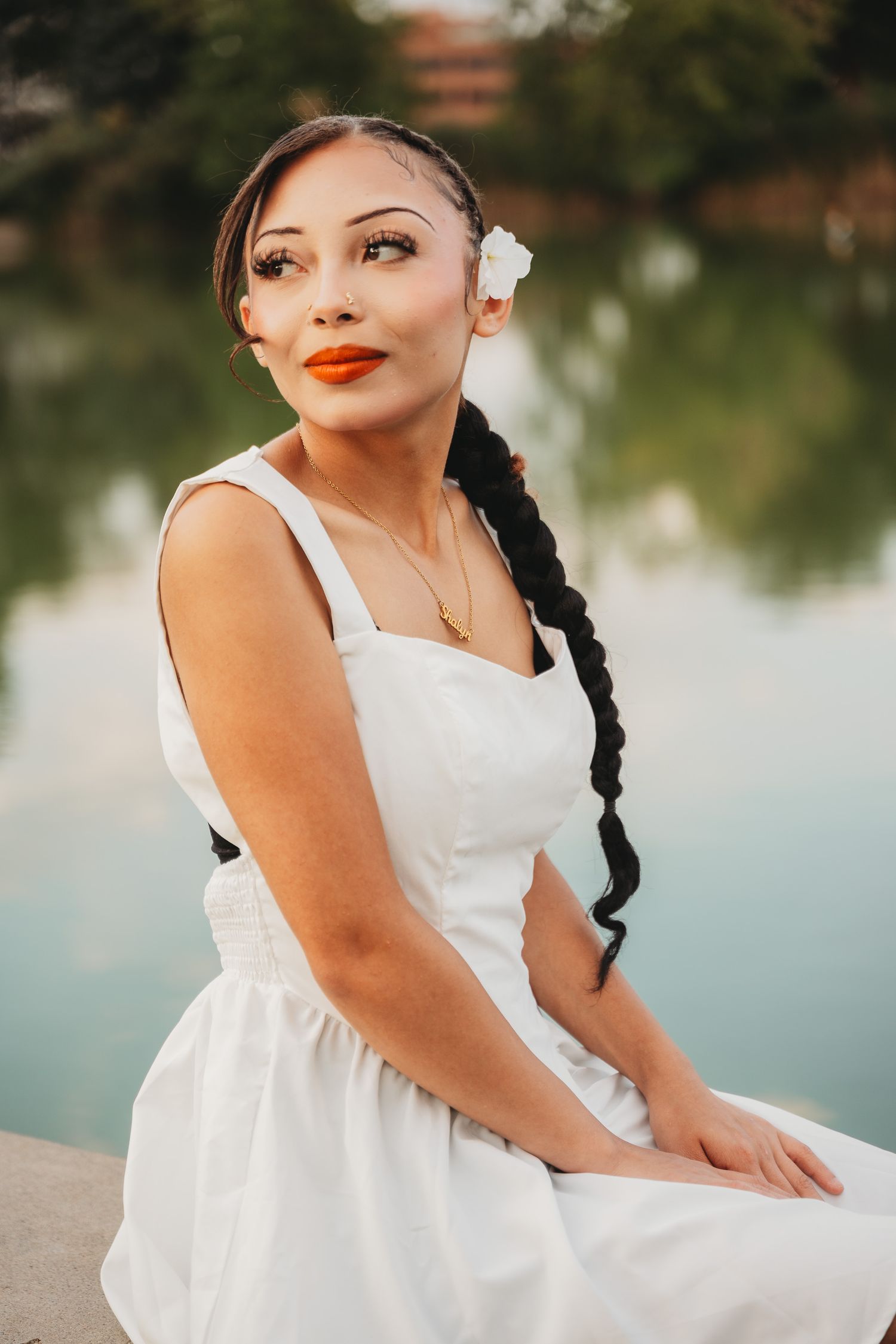 A woman in a flowing white dress sits gracefully by a tranquil lake during a summer photo session.