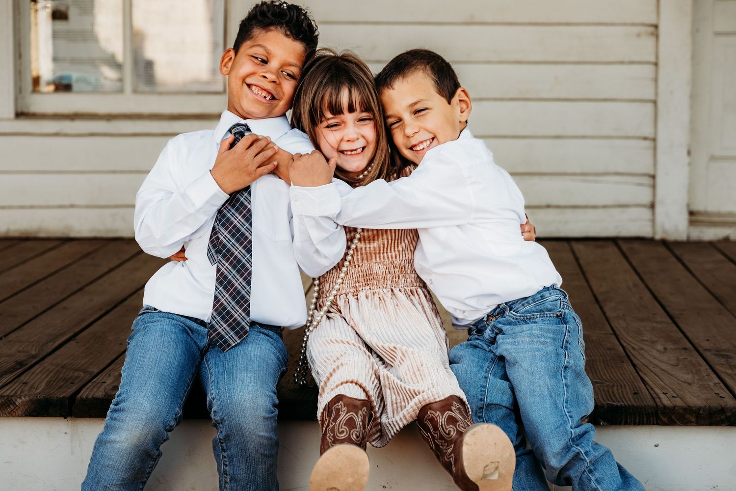 Three children in casual clothing sitting together and laughing on a wooden porch.