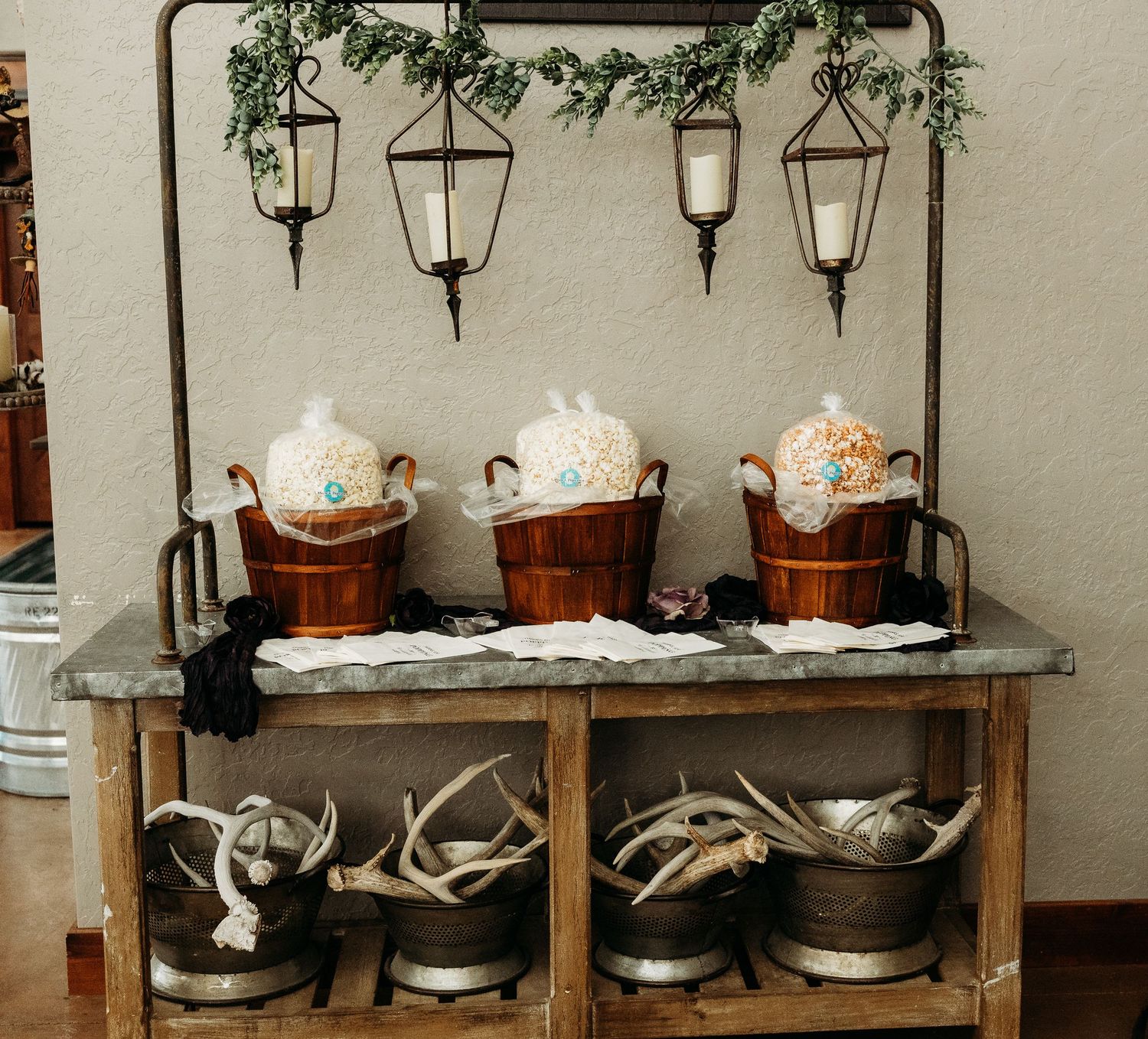 Rustic wedding drink station with copper beverage dispensers, antler decor, and hanging lanterns on a vintage metal table.