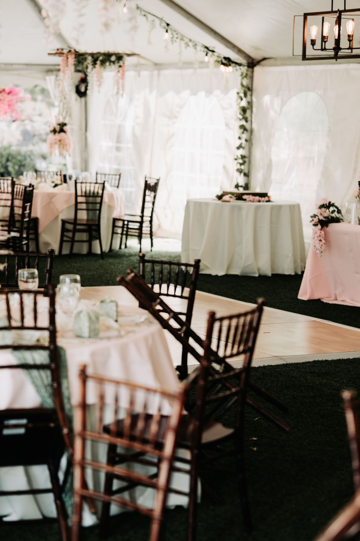 Elegant wedding reception tent with chiavari chairs, pink floral centerpieces, white tablecloths, and a wooden dance floor.