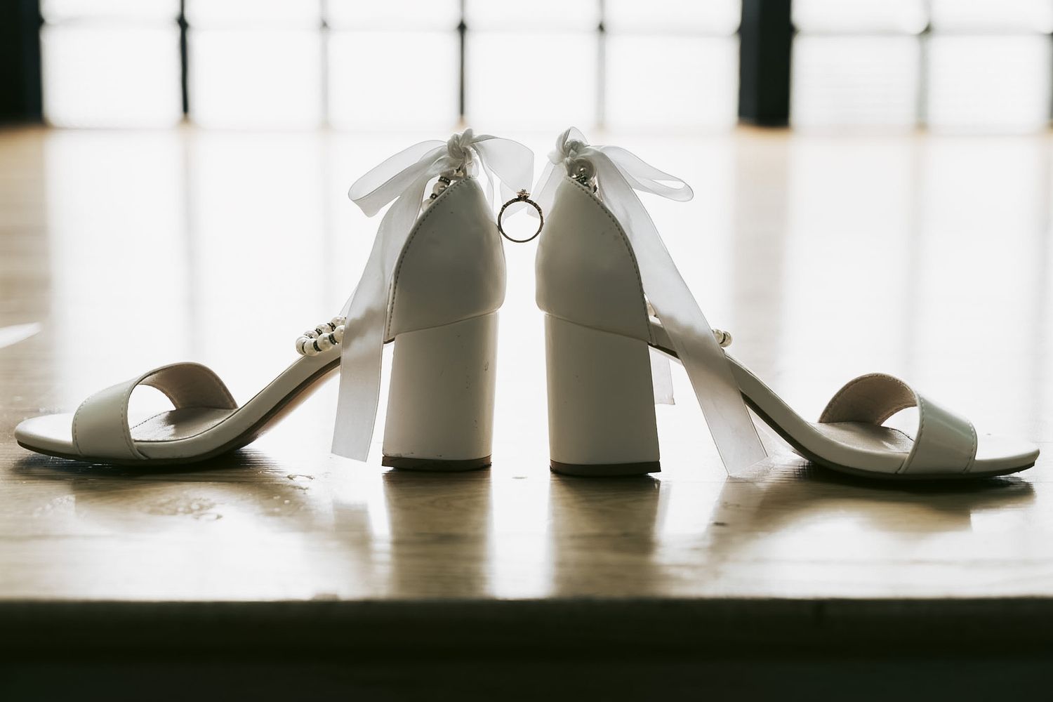 White block heel bridal sandals with bow detail displayed on wooden floor with wedding rings between them.