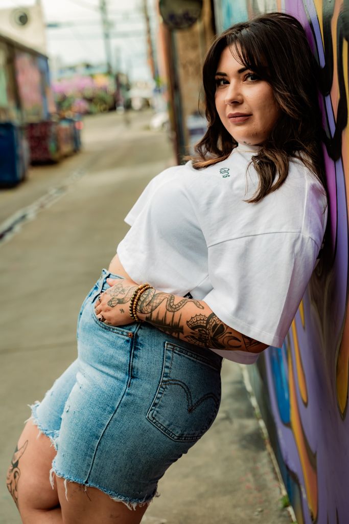 Woman in white crop top and denim shorts posing against urban graffiti wall.