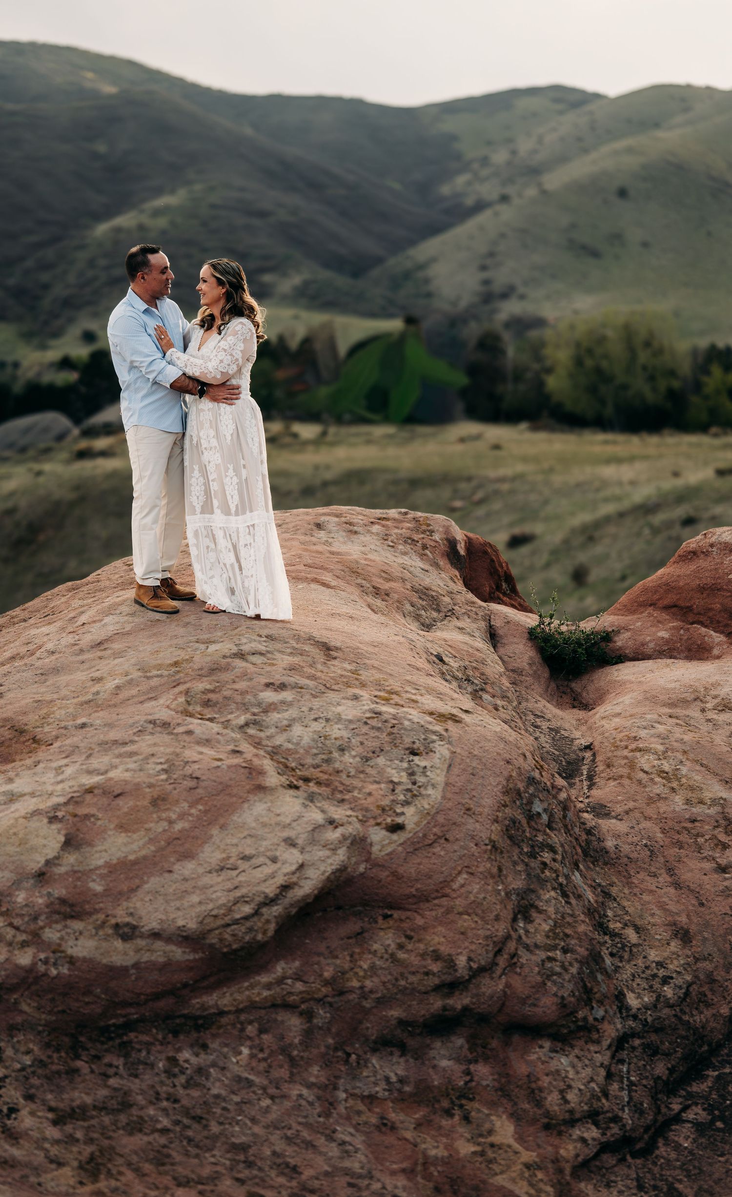 Couple in white attire standing on red rock formation with green rolling hills backdrop during outdoor engagement session.