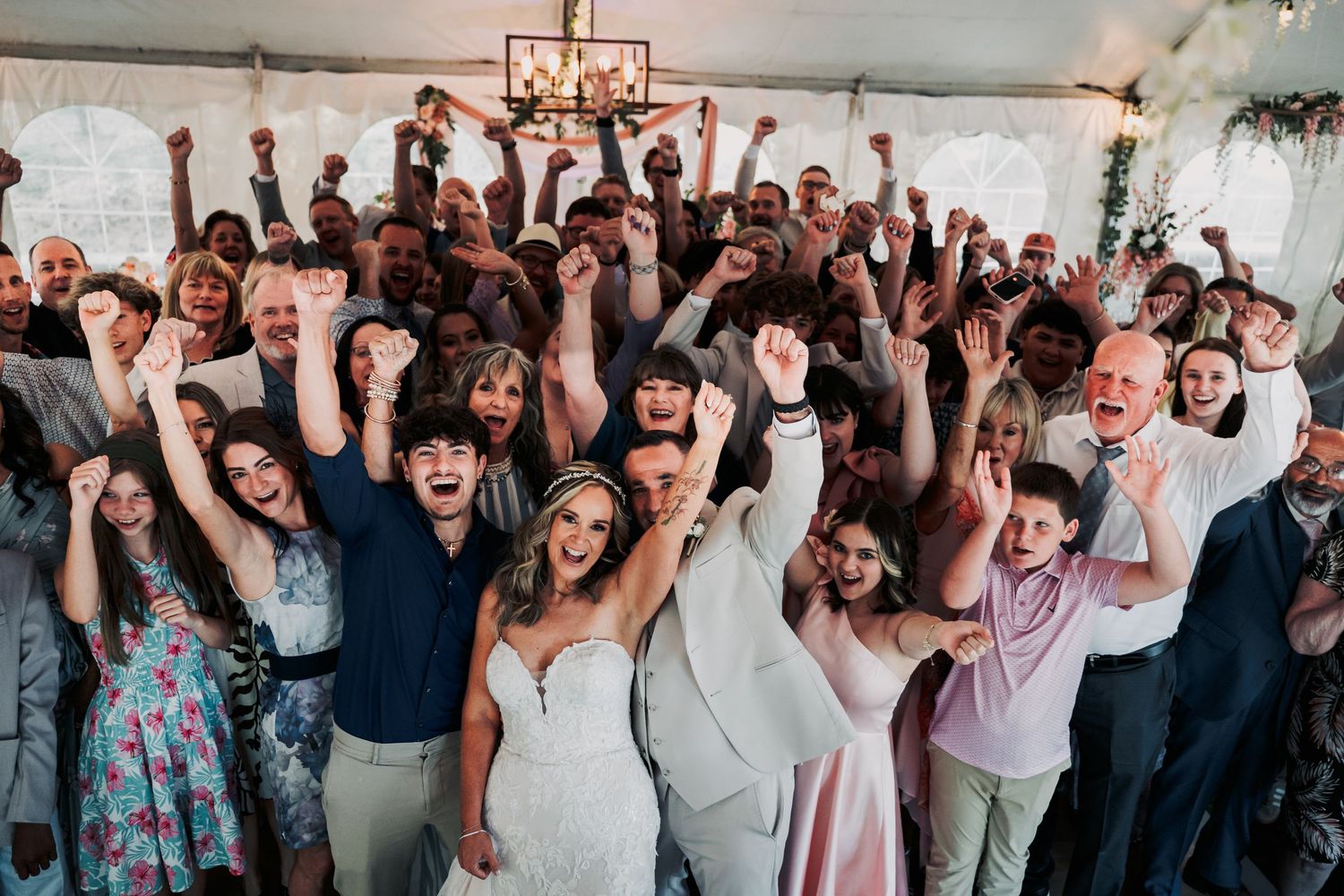 Joyful wedding guests and couple celebrate together with raised arms at a lively indoor reception.