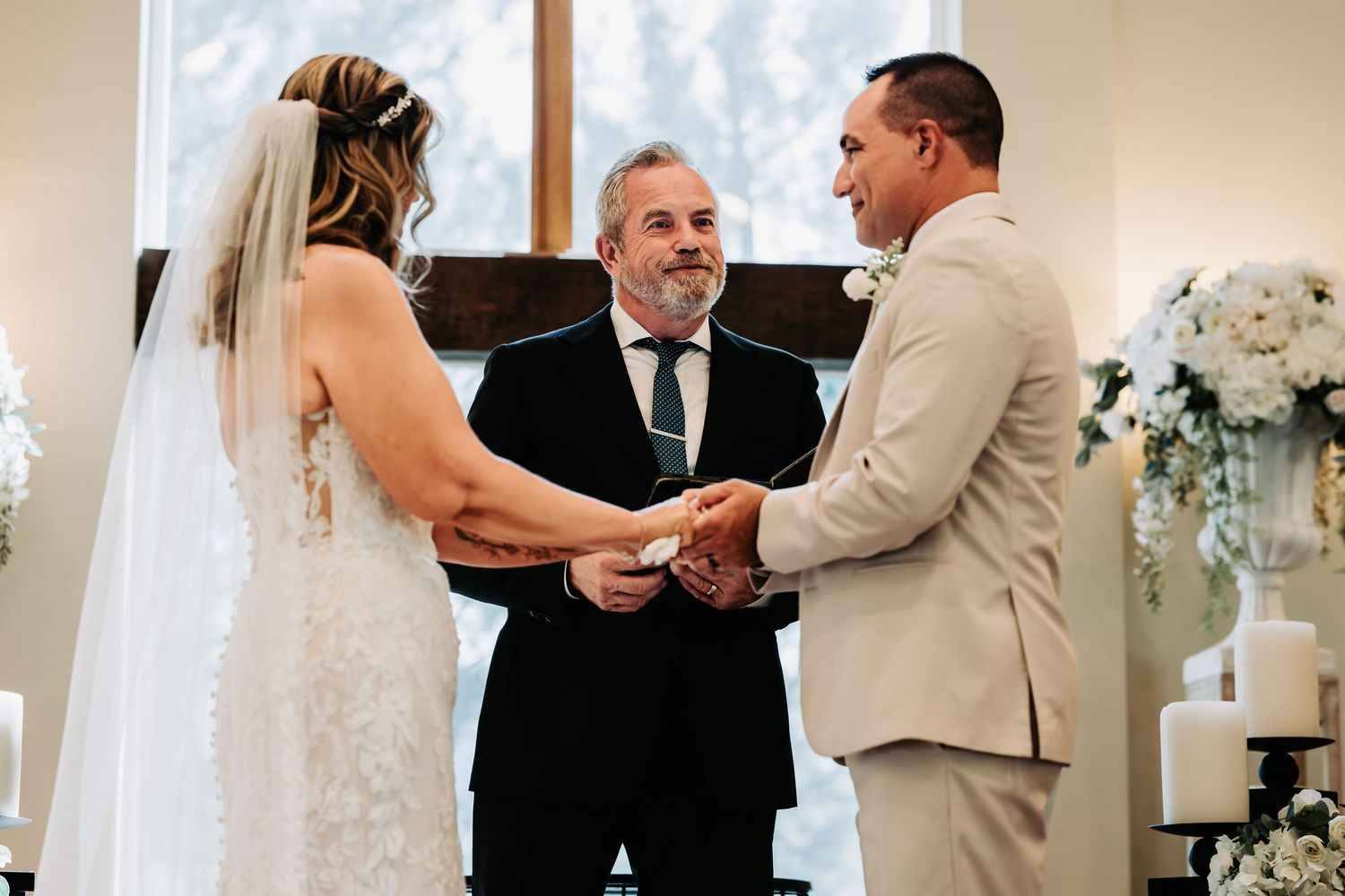 Bride and groom exchange rings during indoor wedding ceremony with officiant and white floral arrangements.