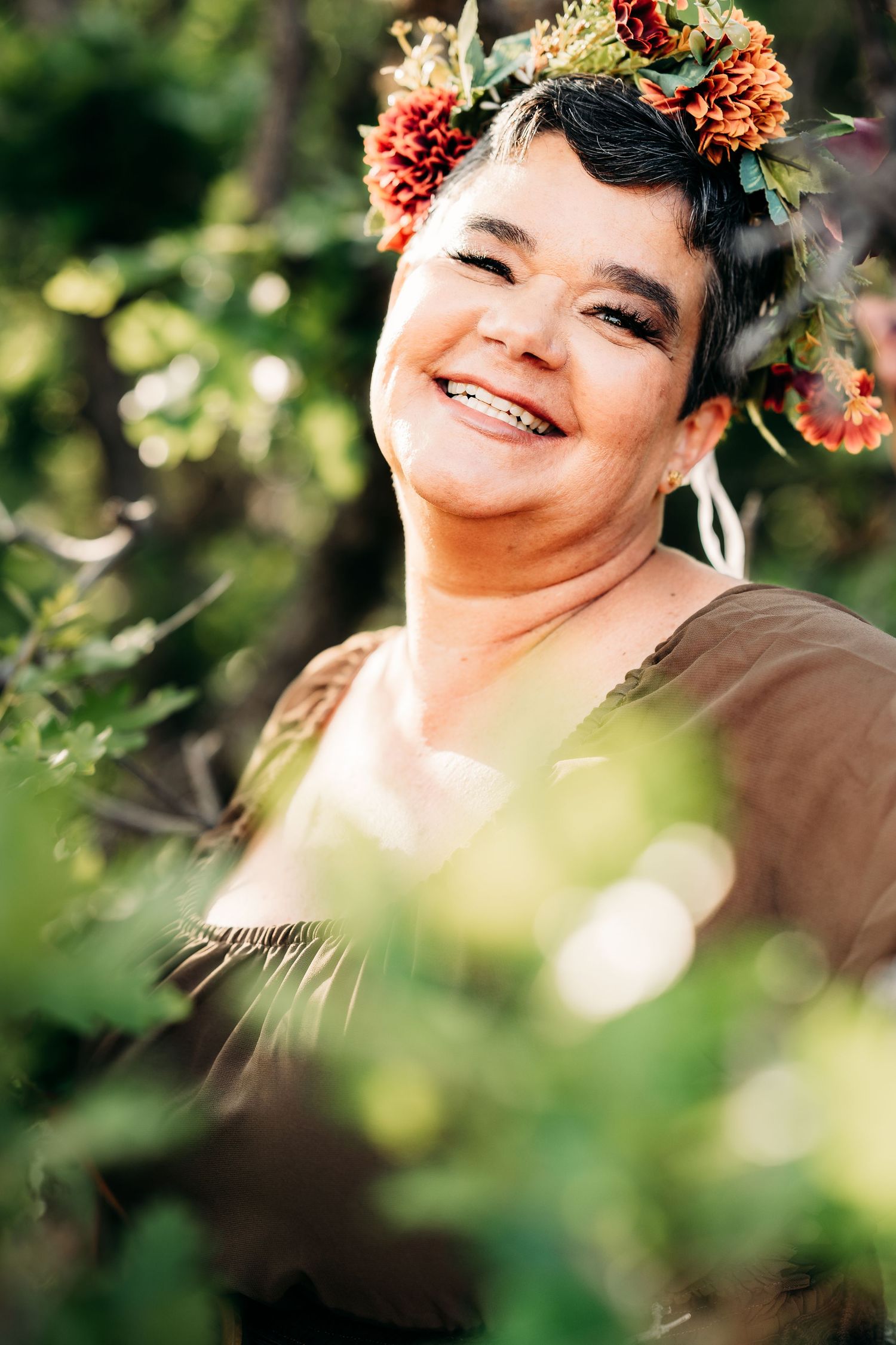 Smiling woman wearing a colorful floral crown surrounded by lush green foliage outdoors.
