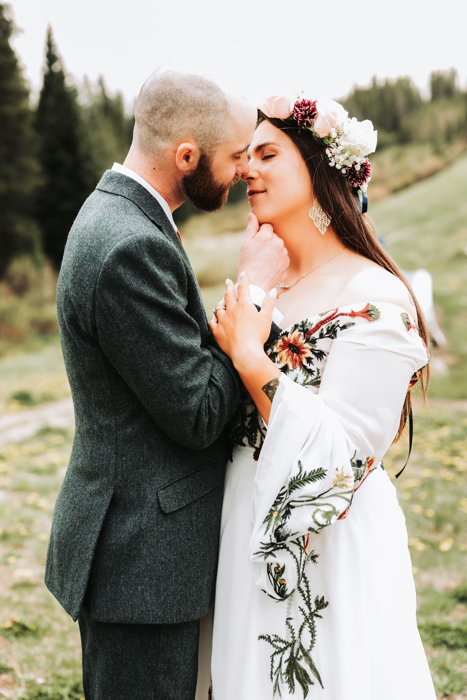 Bride in floral embroidered dress and flower crown shares romantic moment with bearded groom in gray suit outdoors.
