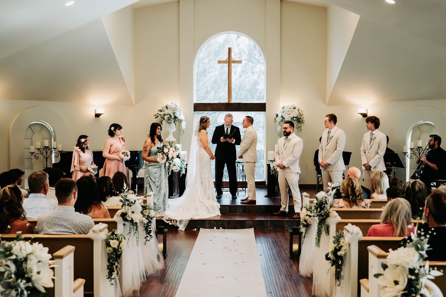 Elegant wedding ceremony in a white chapel with cross, bride and groom at altar with wedding party and floral arrangements.