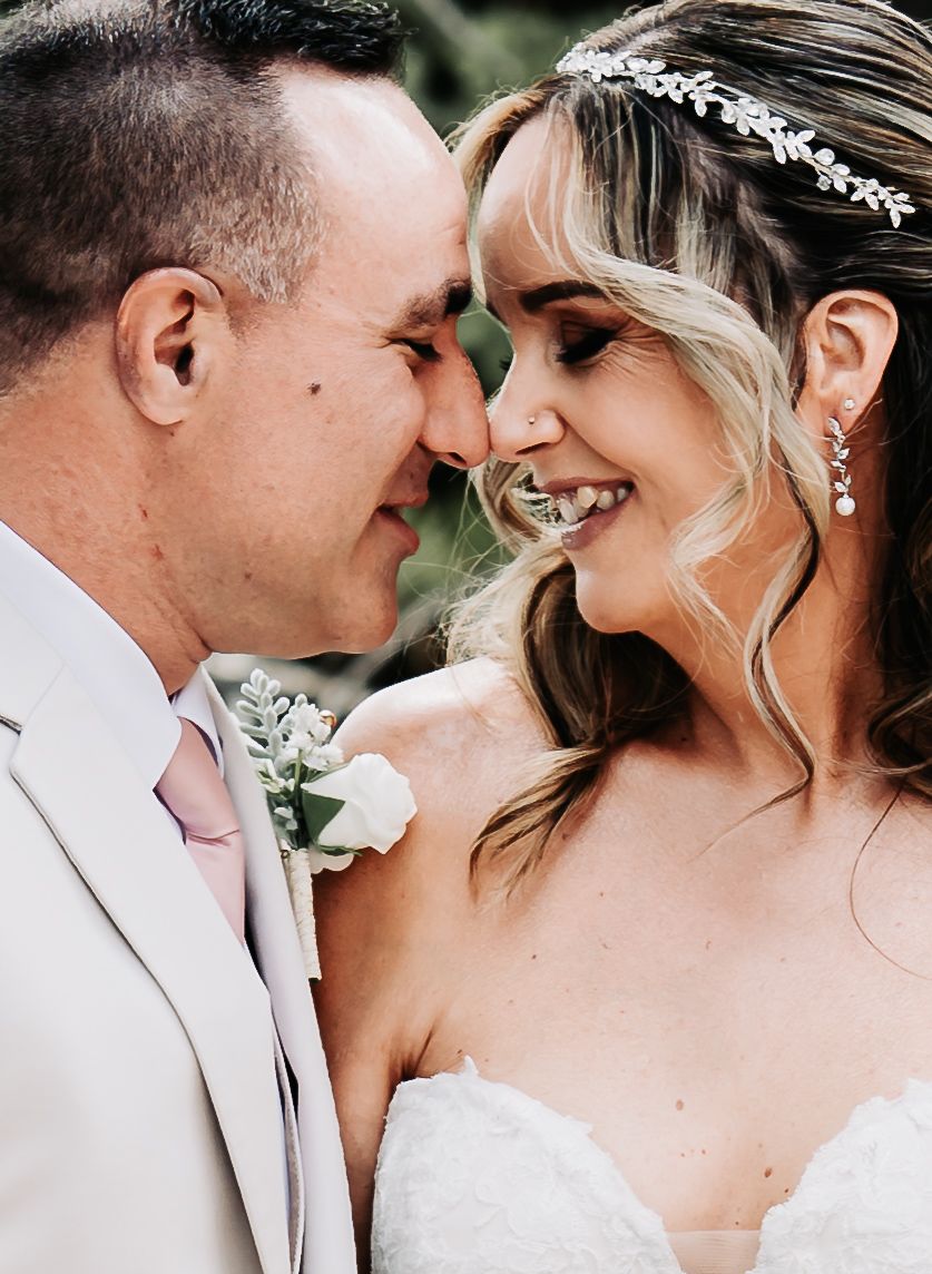A smiling bride in a white gown and floral headpiece shares a romantic moment with her groom in a light suit.