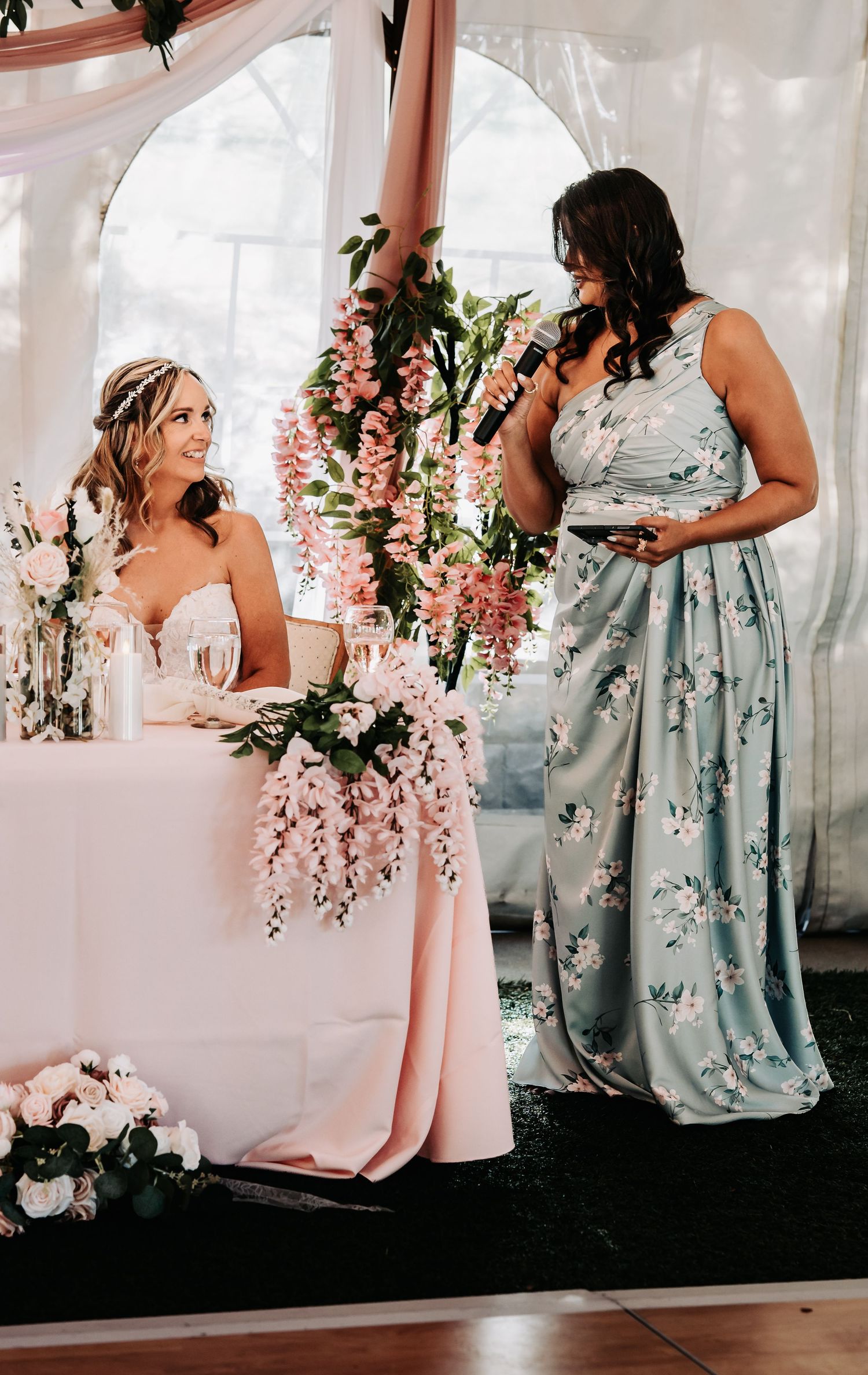 Bride smiles at bridesmaid giving a speech at a floral-decorated wedding reception table with pink flowers.