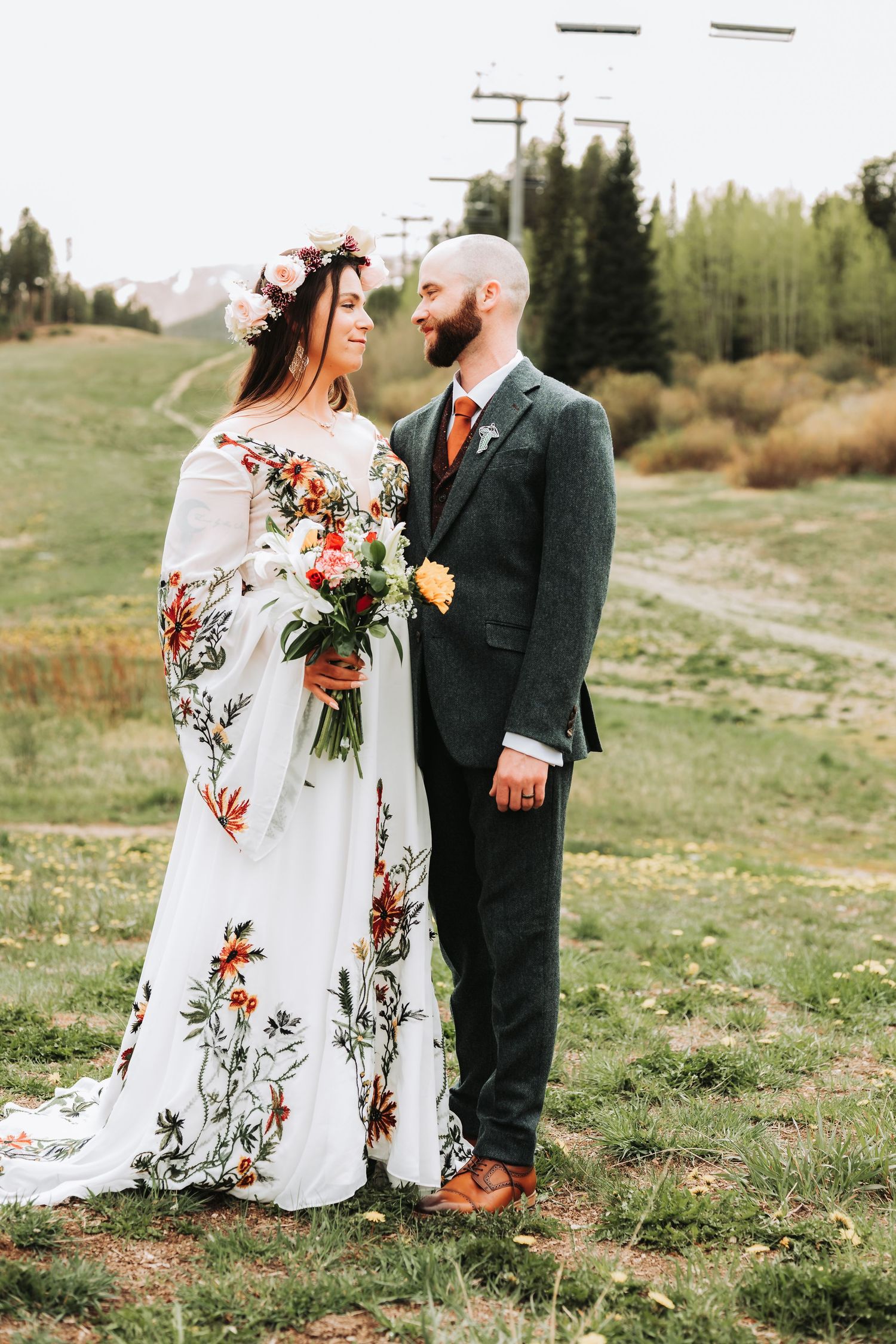 Bride in floral embroidered dress with flower crown holds bouquet beside groom in gray suit on green hillside.