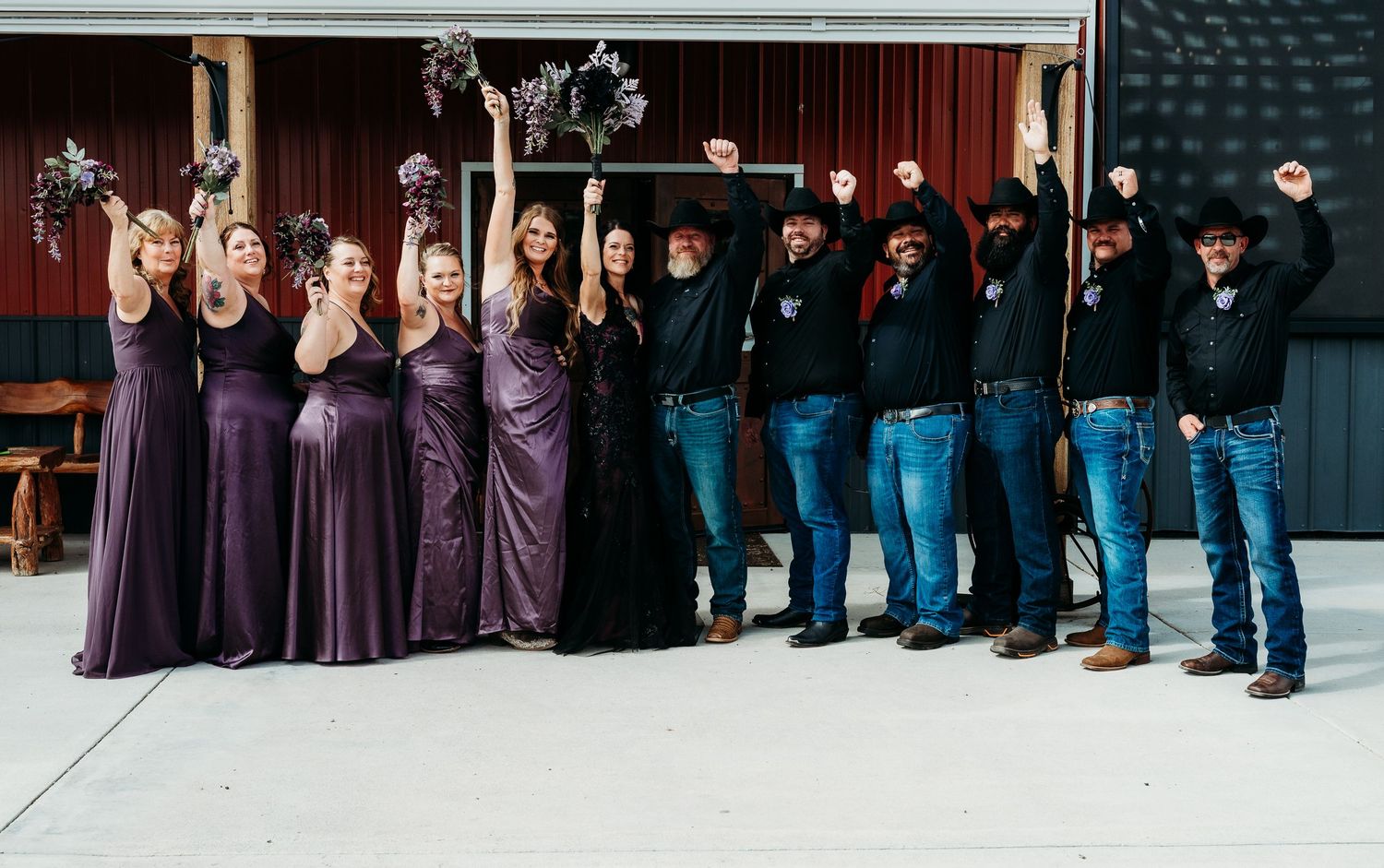 Wedding party celebrating with raised fists, bridesmaids in purple gowns and groomsmen in black shirts and jeans.